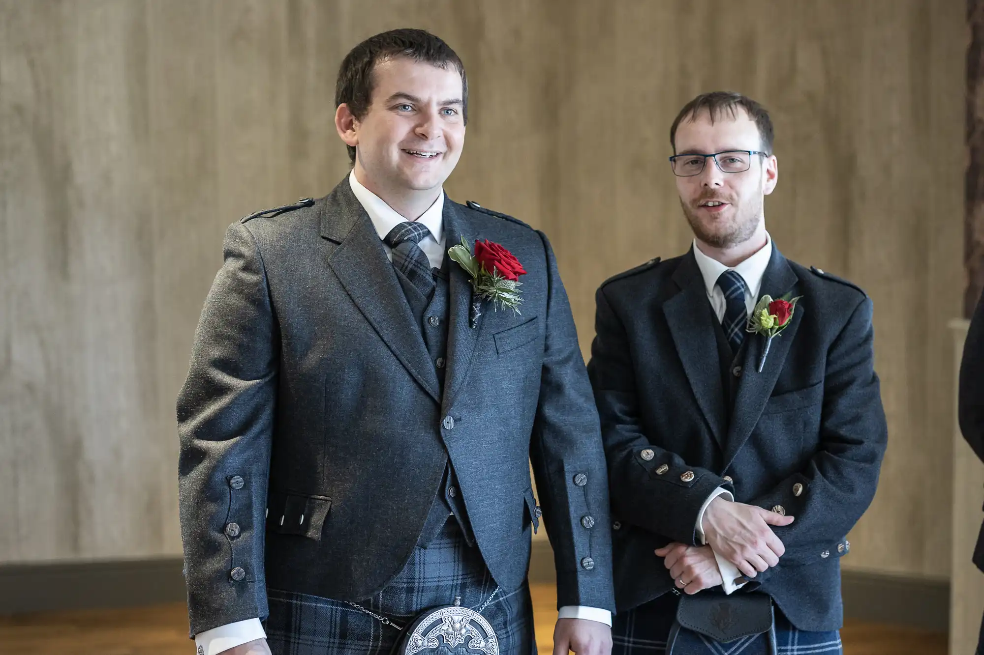 Two men wearing grey Scottish formal attire with kilts and jackets stand indoors. Both have red rose buttonholes. The man on the left smiles broadly, while the man on the right, with glasses, stands with arms folded, facing forward. The background is neutral.