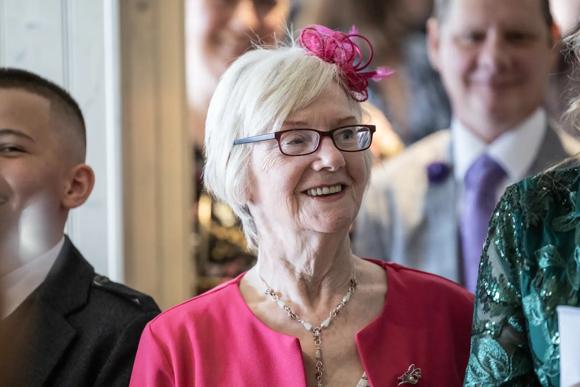 An elderly woman with short white hair, glasses, and a bright pink fascinator smiles. She wears a matching pink jacket and a delicate necklace. People in formal attire appear blurred in the background, suggesting a celebratory event.
