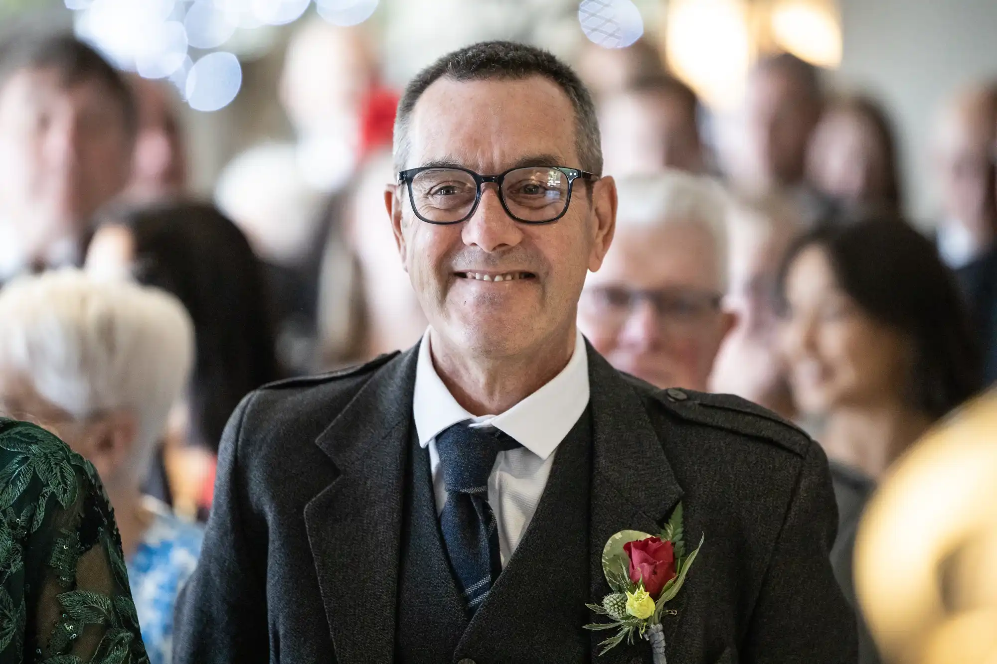 A smiling man with glasses wears a dark suit jacket, tie, and a buttonhole with a red rose. He stands indoors among a blurred crowd, suggesting a formal event or celebration. Soft lighting and fairy lights are visible in the background.