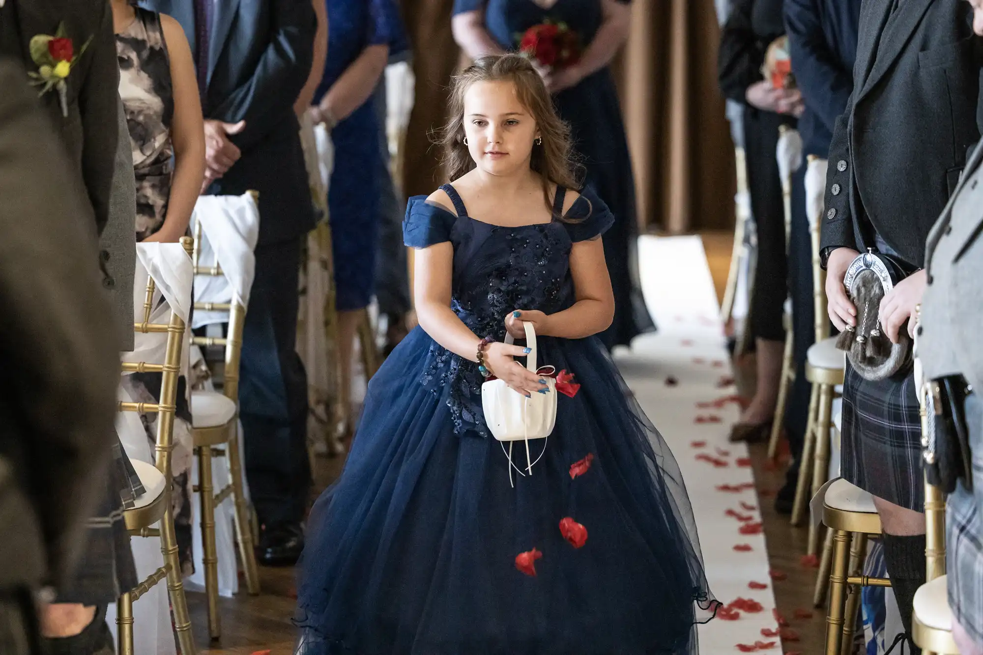 A young girl in a navy blue dress walks down a white aisle, scattering red rose petals from a white basket. She is at a formal event, likely a wedding, surrounded by seated guests in formal attire. Rose petals are spread on the aisle floor.