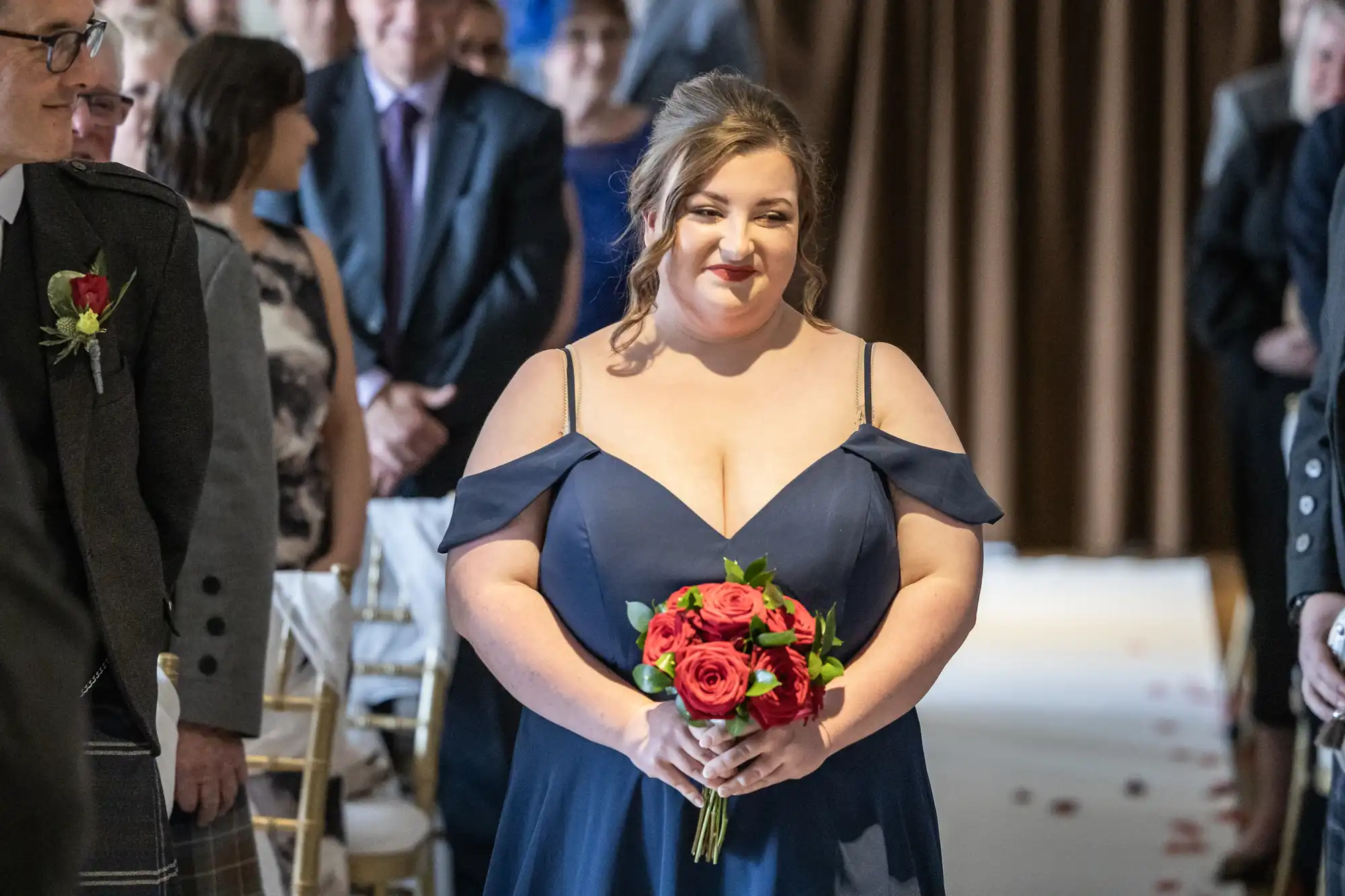 A woman in a navy blue, off-the-shoulder dress holds a bouquet of red and pink roses. She is smiling and walking down an aisle lined with seated guests, in a well-lit indoor setting, possibly at a wedding or formal event.