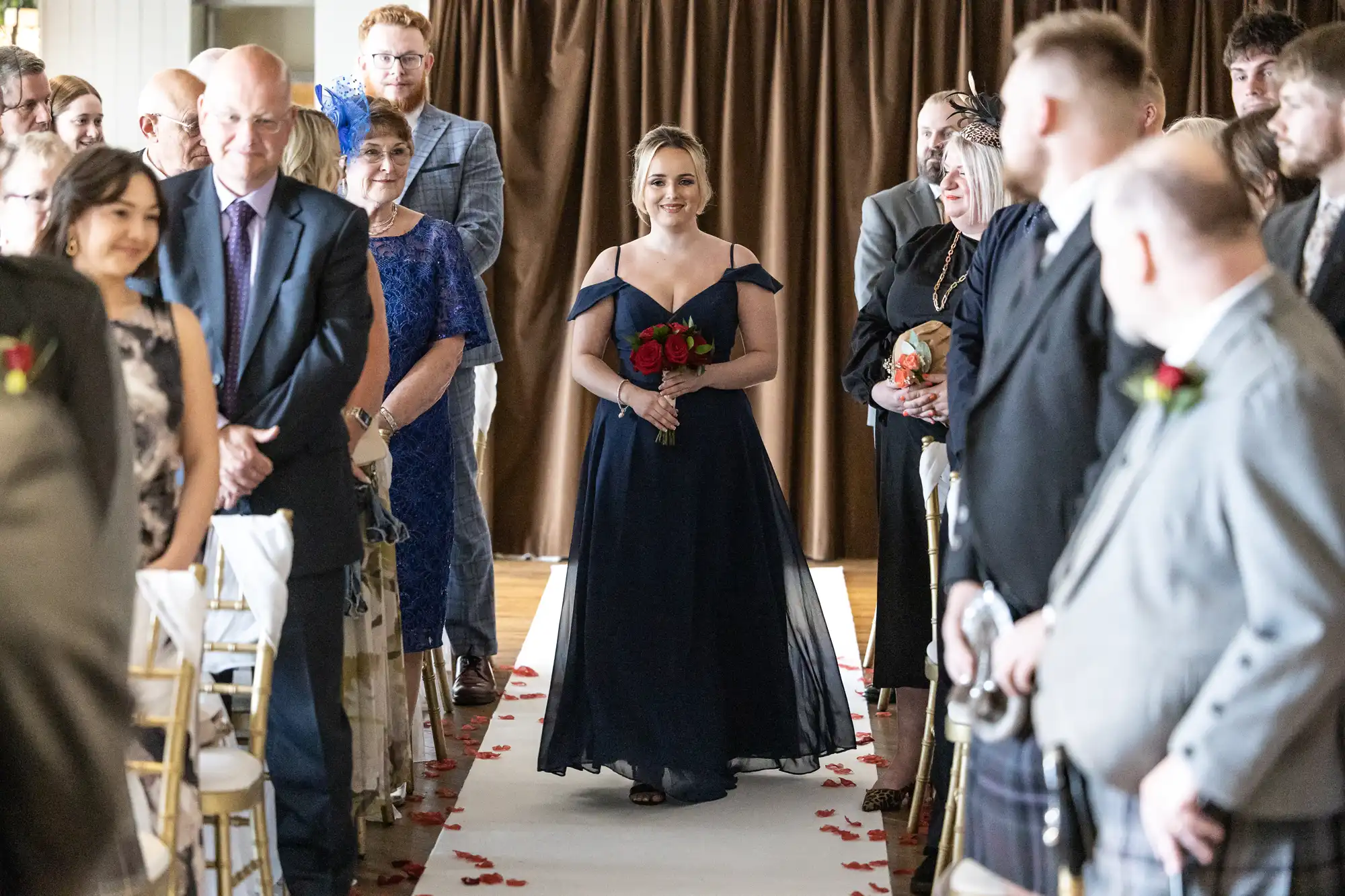 A woman in a navy blue gown holding red roses walks down an aisle lined with seated guests on both sides. The aisle is covered in white fabric with scattered red petals, and brown curtains hang in the background. Guests watch her, some smiling.