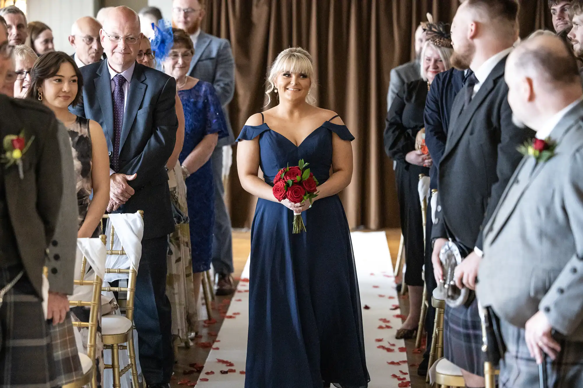 A bridesmaid in a navy blue off-shoulder dress walks down a petal-lined aisle, smiling and holding a red bouquet, whilst formally dressed wedding guests seated on either side watch her procession.