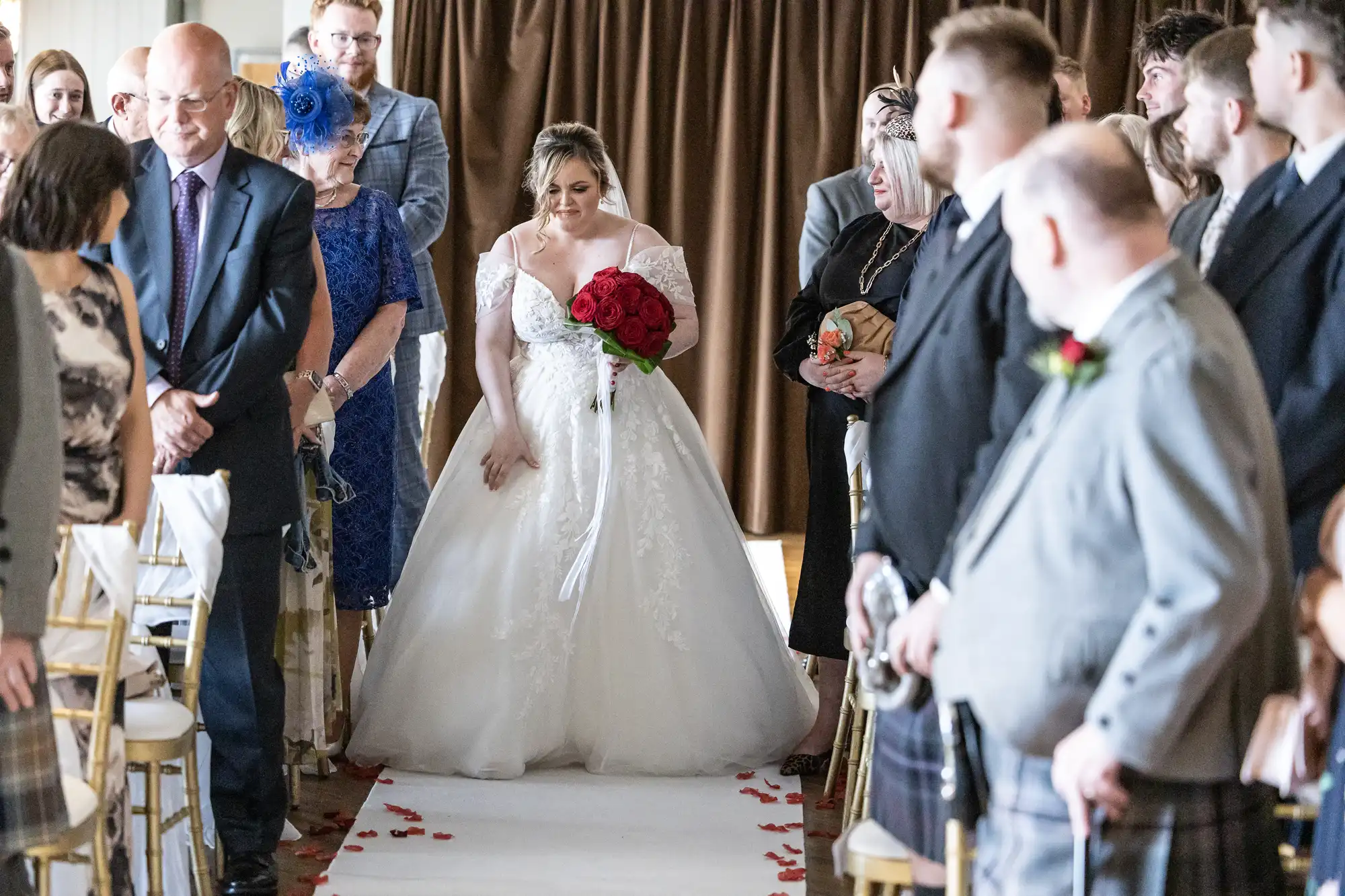 A bride in a white dress holding red roses walks down an aisle covered with rose petals, smiling and looking down. Guests in formal attire stand on either side of the aisle, watching her. Brown curtains are visible in the background.