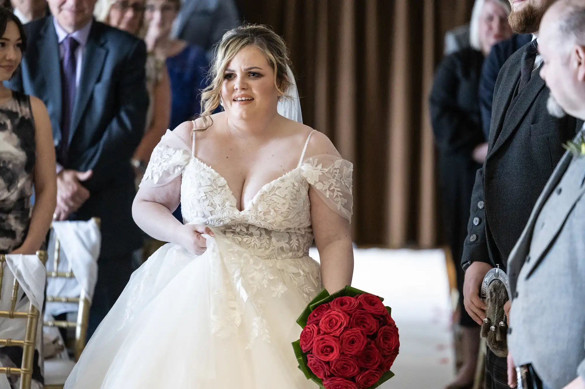 A bride in a white lace wedding dress holding a red rose bouquet walks down the aisle, surrounded by seated guests in formal attire, with a man in a suit standing nearby and a brown curtain in the background.