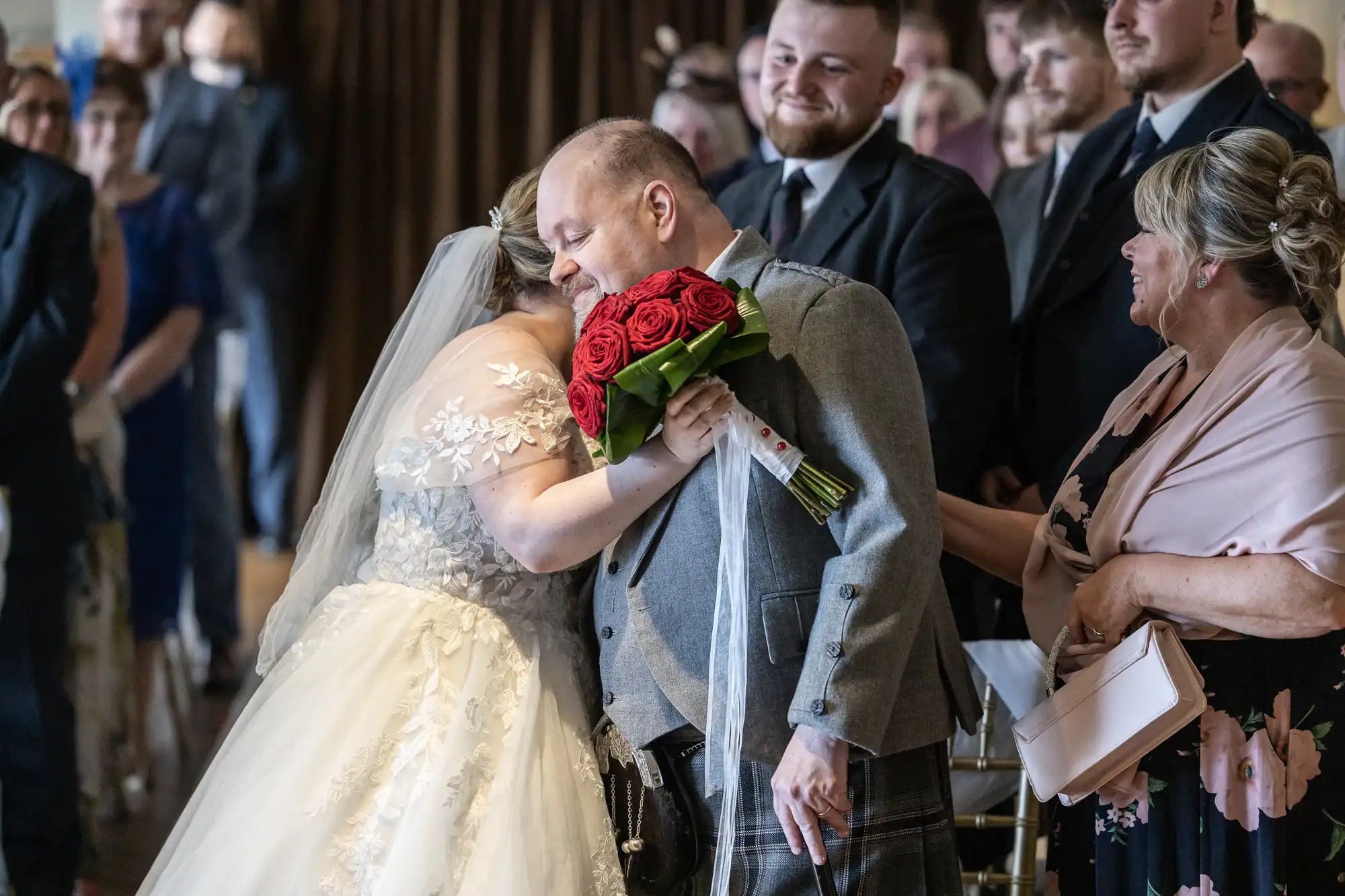 A bride in a white gown hugs an older man in a grey suit and tartan kilt, holding red roses. People dressed formally watch and smile in the background, seated in rows, suggesting a heartfelt moment during a wedding ceremony.