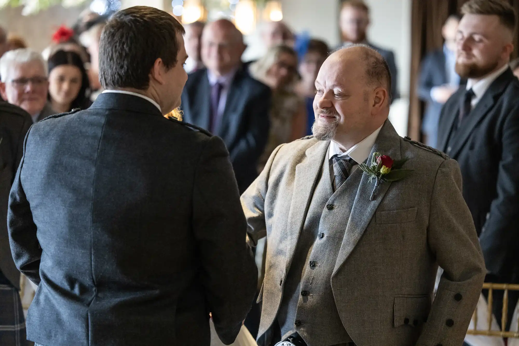 Two men in grey suits shake hands and smile warmly during a formal event, surrounded by seated and standing guests in the background. One man wears a buttonhole with a red rose, and both are dressed in traditional Scottish attire.