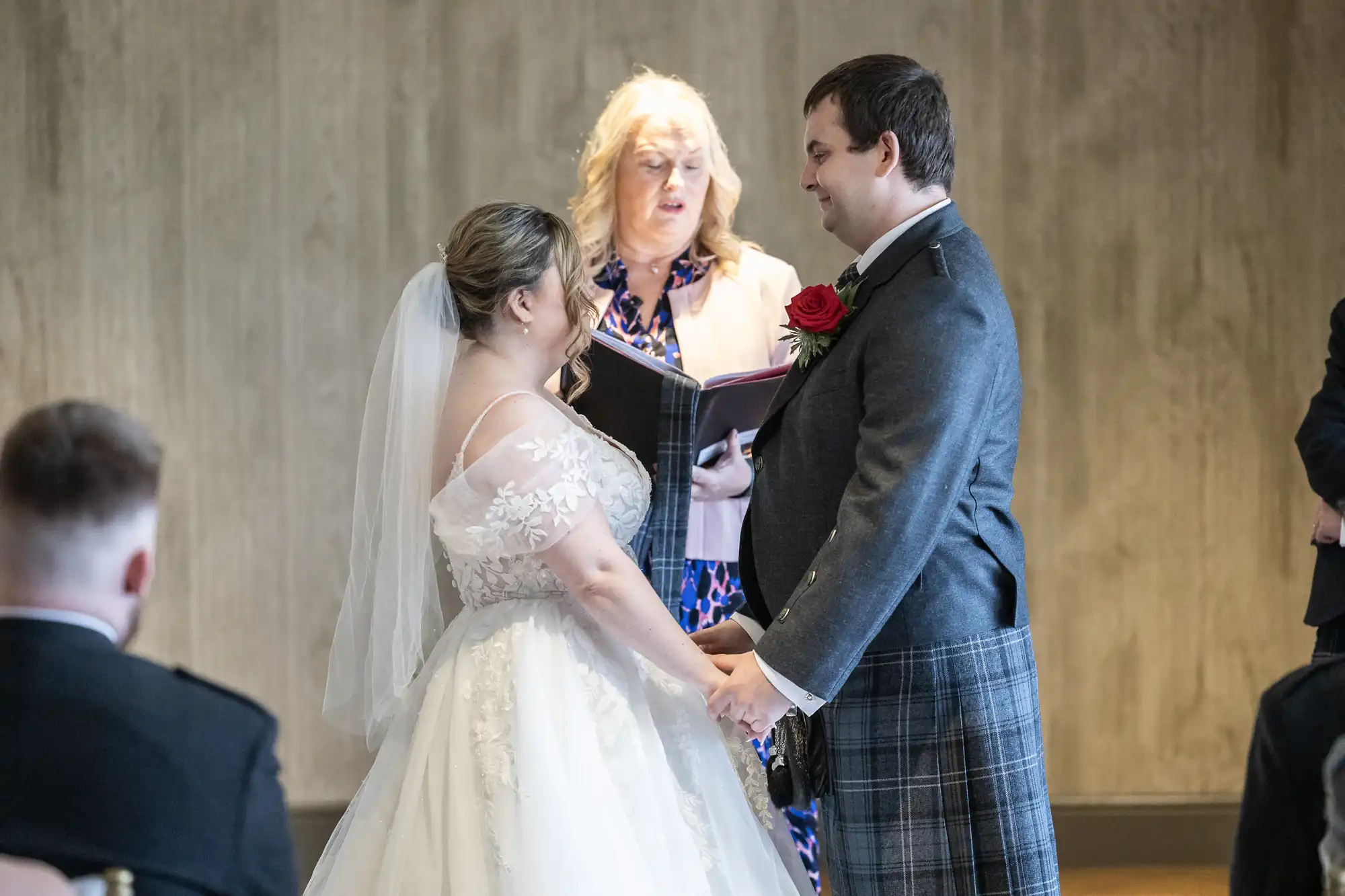 A bride and groom stand holding hands and smiling at each other during their wedding ceremony. The bride wears a white dress with lace details and a veil. The groom wears a grey jacket and checked kilt. An officiant stands behind them, reading from a book.