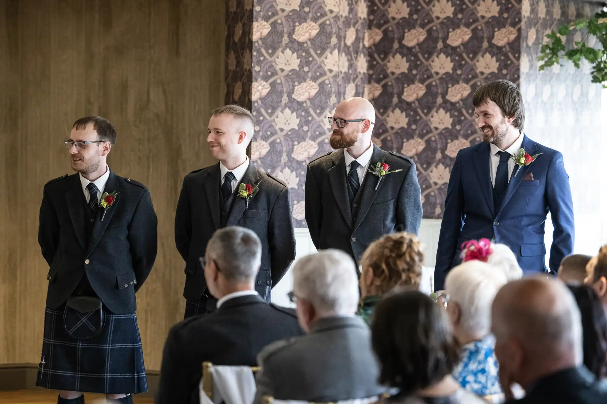 Four men stand at the front of a wedding ceremony. Three wear traditional Scottish kilts and jackets with buttonholes, while one wears a blue suit. They smile and look forward as seated guests watch. The room has patterned wallpaper and flower decorations.