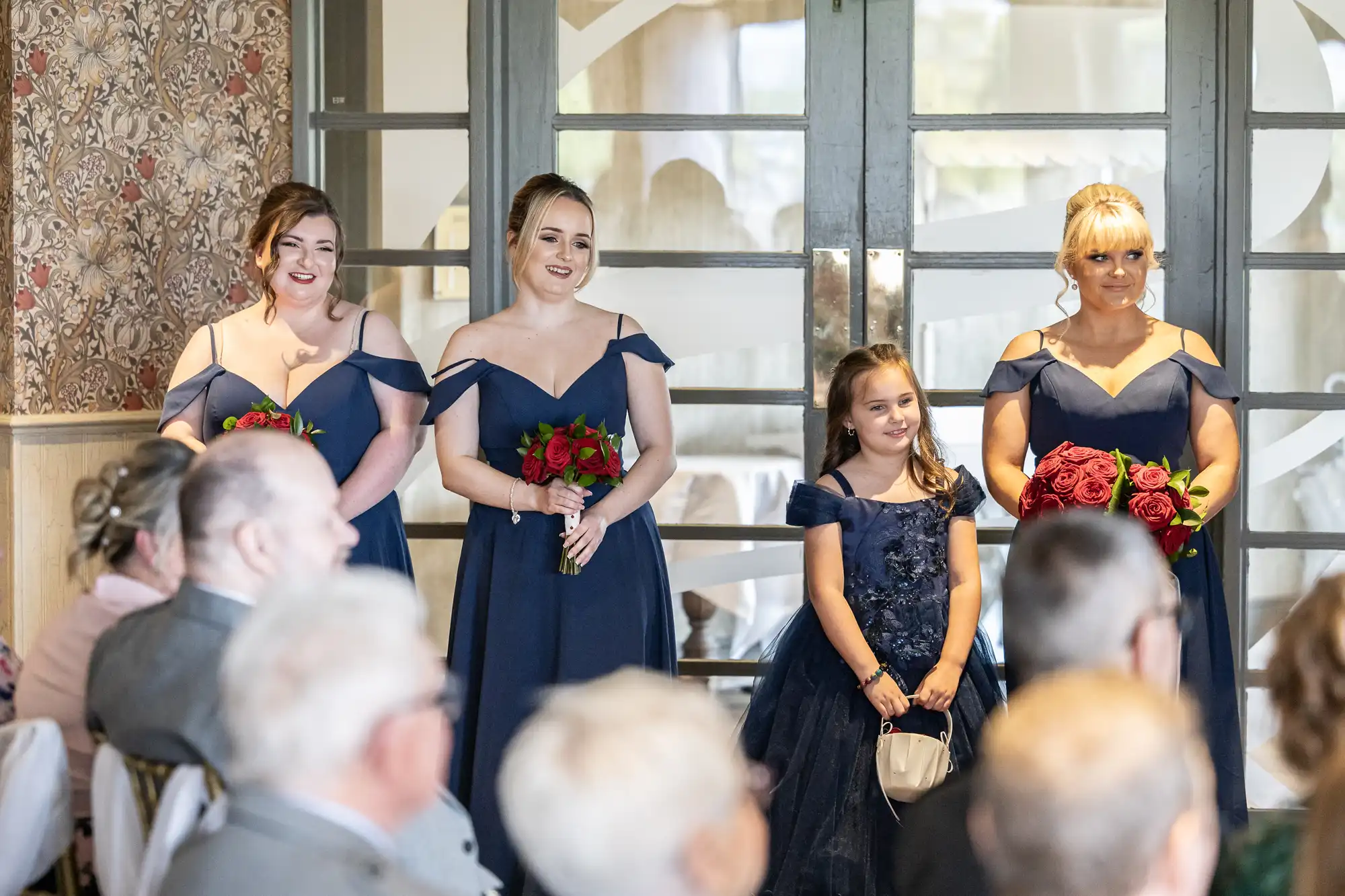 Three bridesmaids in matching navy blue dresses hold red bouquets and smile, standing beside a young girl in a navy dress holding a small bag. Several seated guests face them in a well-lit room with patterned walls and glass doors.