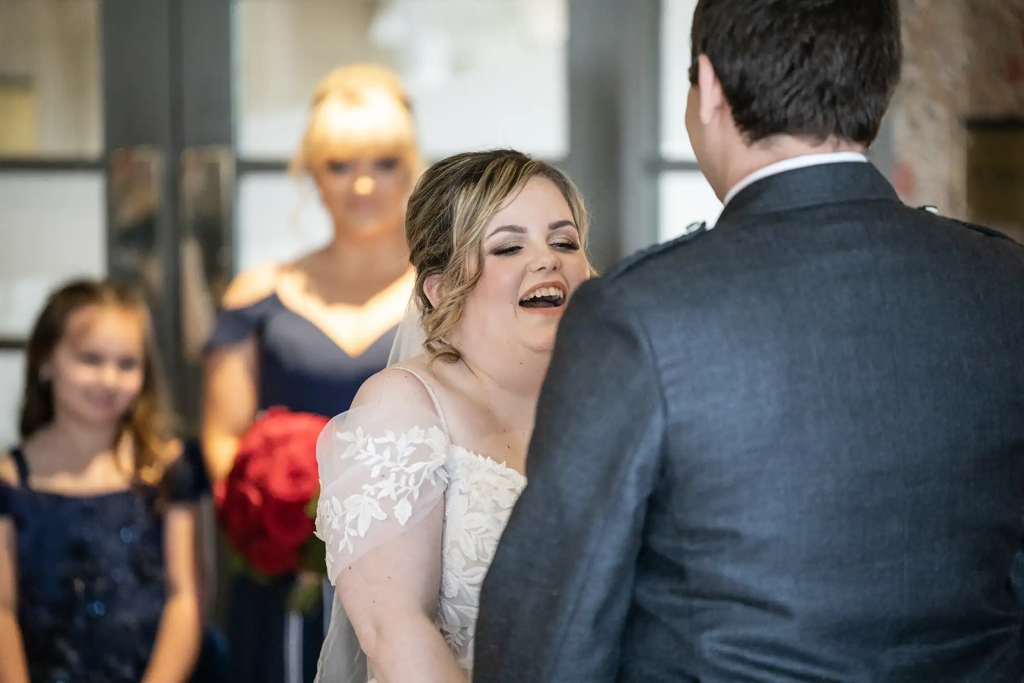 A bride in a white dress laughs joyfully whilst facing her groom, who wears a grey suit. Two bridesmaids in navy dresses, one adult and one child, stand in the background. The setting appears light and festive, suggesting a wedding ceremony.