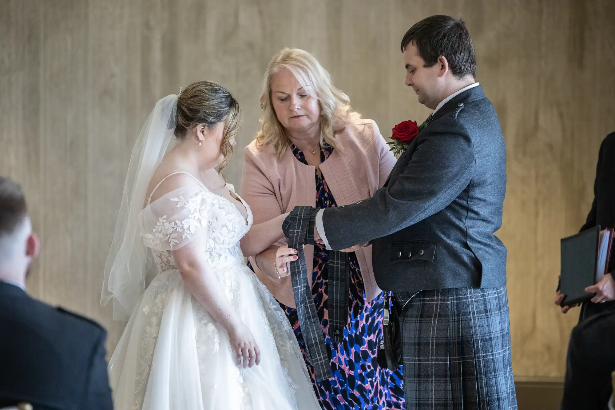 A bride in a white dress and veil and a groom in a kilt stand facing each other. A blonde woman in a pink jacket wraps a tartan cloth around their hands in a handfasting ceremony. The setting is indoors with a neutral background.