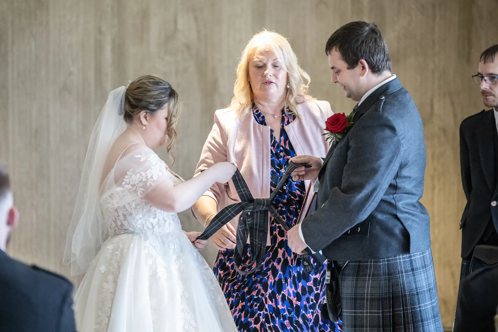 A bride in a white gown and veil and a groom in a dark jacket and kilt hold a tartan fabric together during a wedding ceremony, guided by a woman in a pink jacket and patterned dress, with another man watching in the background.