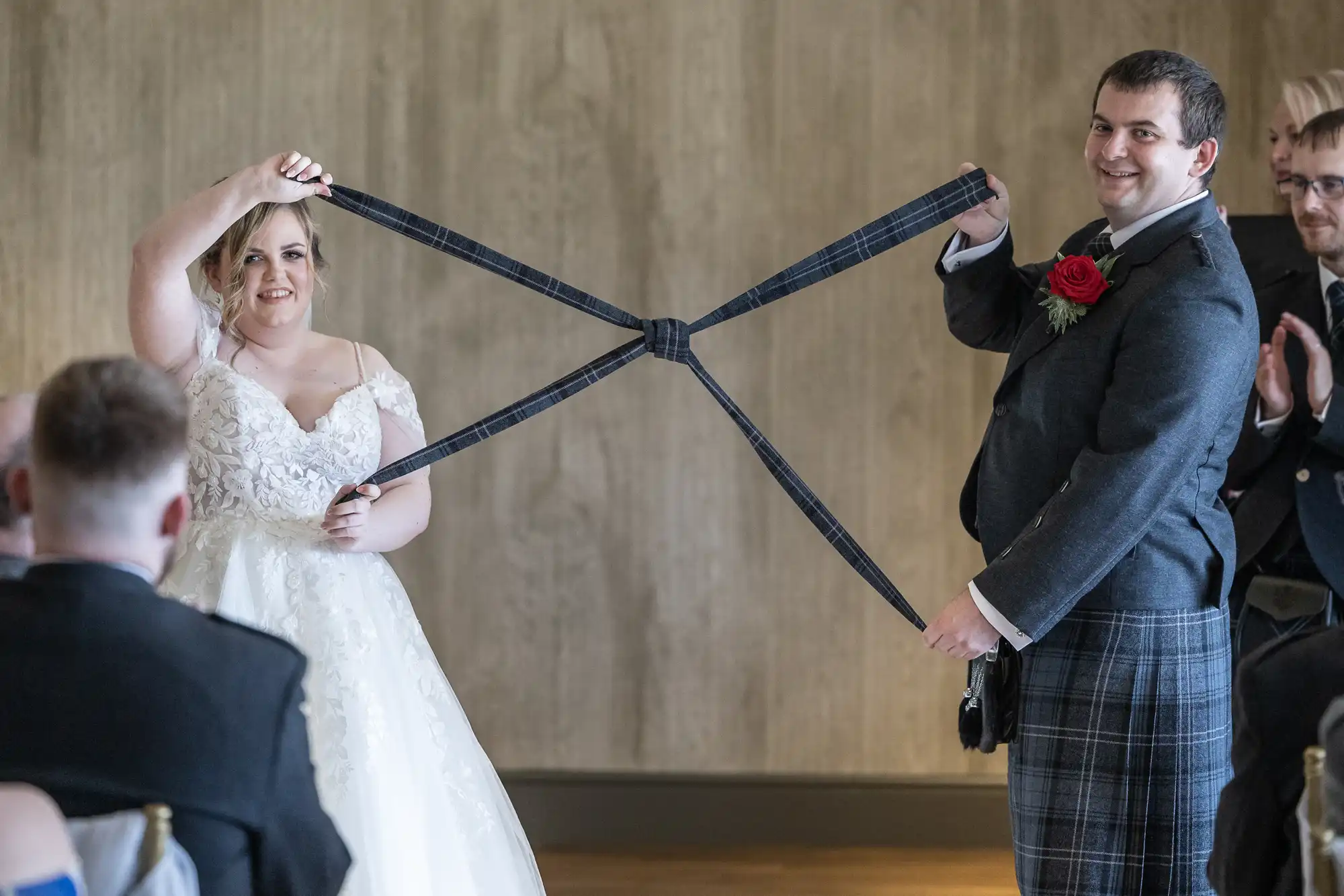 A bride in a white gown and a groom in a kilt hold a tartan cloth together, forming a cross between them, during a wedding ceremony with guests seated around them. Both are smiling, standing in front of a plain, light brown wall.