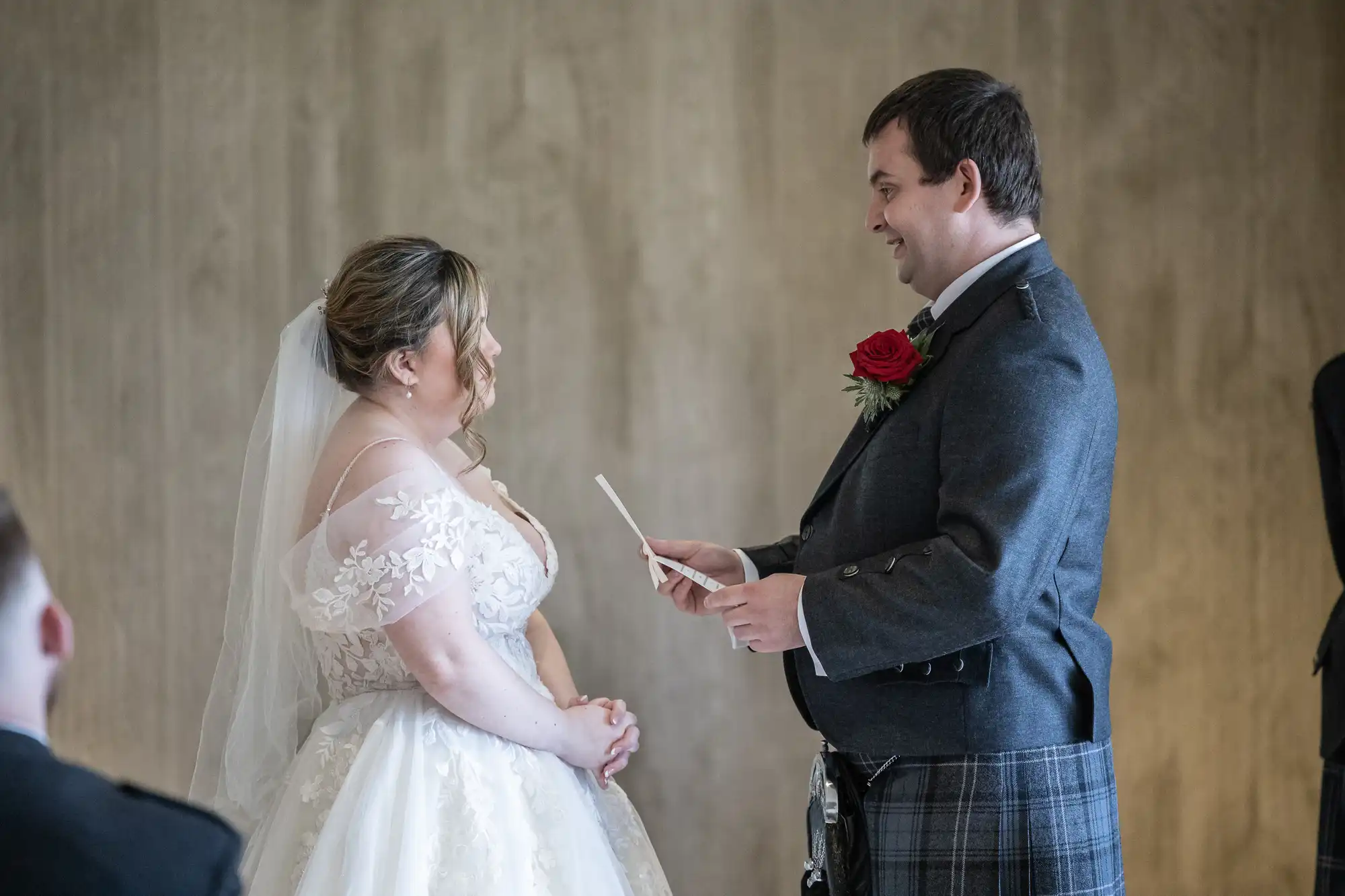 A bride in a white, floral lace wedding dress and veil faces a groom in a dark jacket and tartan kilt. The groom smiles whilst reading from a card. They stand indoors against a plain beige background, sharing a moment during their wedding ceremony.