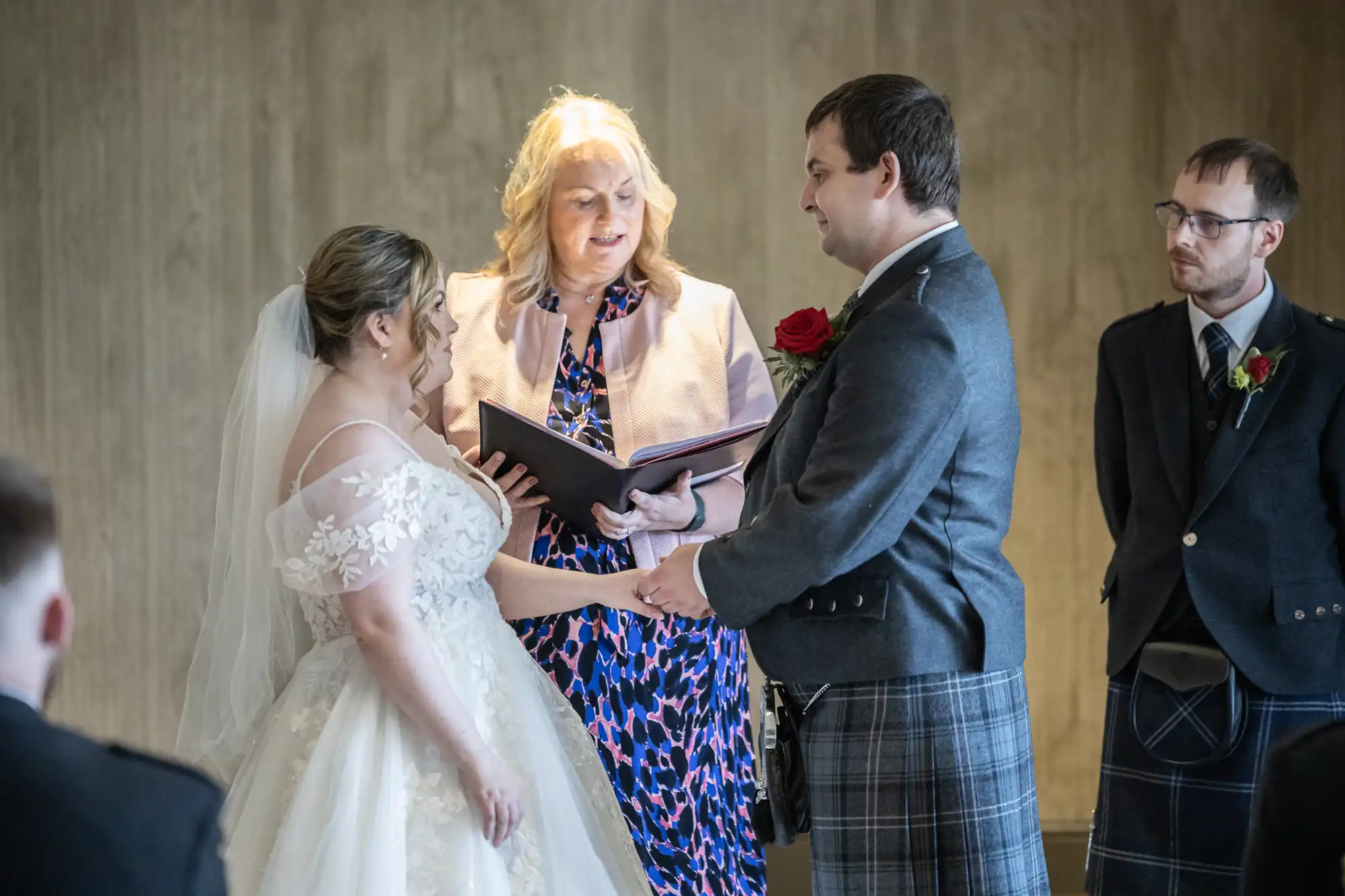 A bride and groom stand facing each other, holding hands during a wedding ceremony. An officiant stands between them, reading from a book. Another man in a dark suit with a red rose on his lapel stands to the side, watching. The background is neutral and softly lit.
