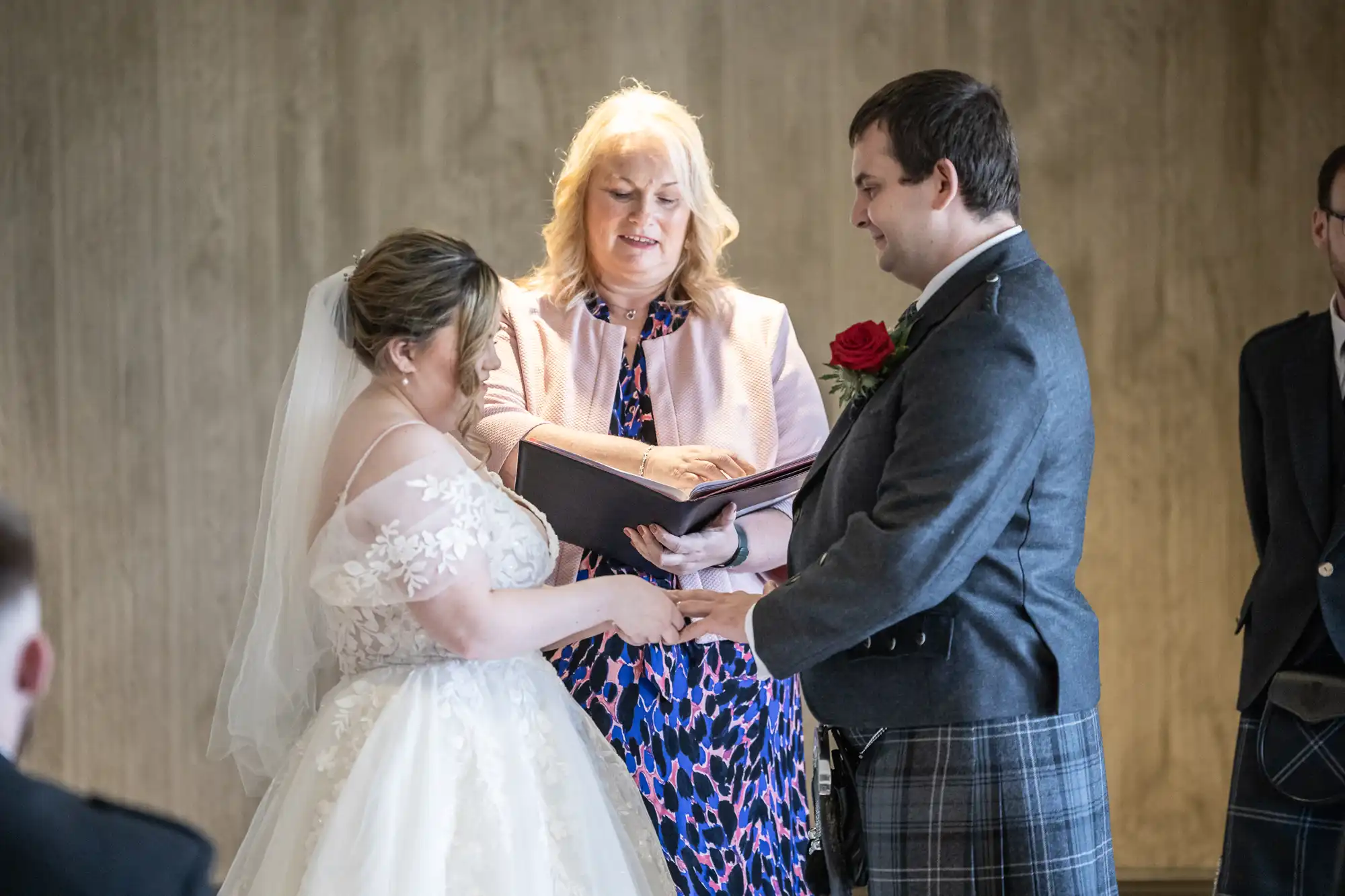 A bride and groom stand facing each other, holding hands and smiling, as an officiant in a light pink jacket reads from a book. The bride wears a white dress and veil; the groom wears a kilt with a red rose buttonhole. The background is softly lit and neutral.