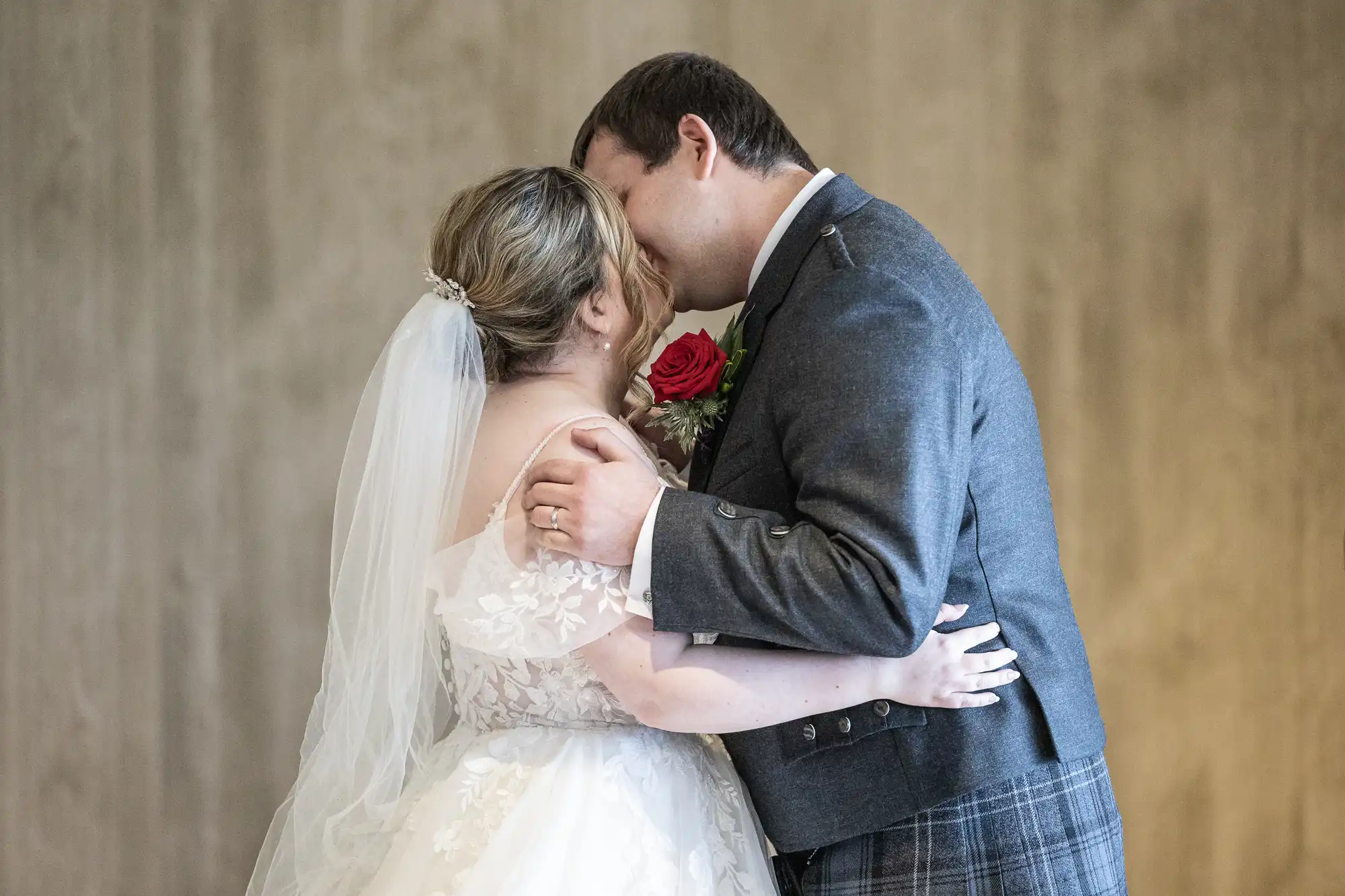 A bride in a white dress and veil embraces and kisses a groom in a grey jacket and checked kilt. The groom holds a red rose buttonhole. Both appear joyful, standing close against a plain, beige background.