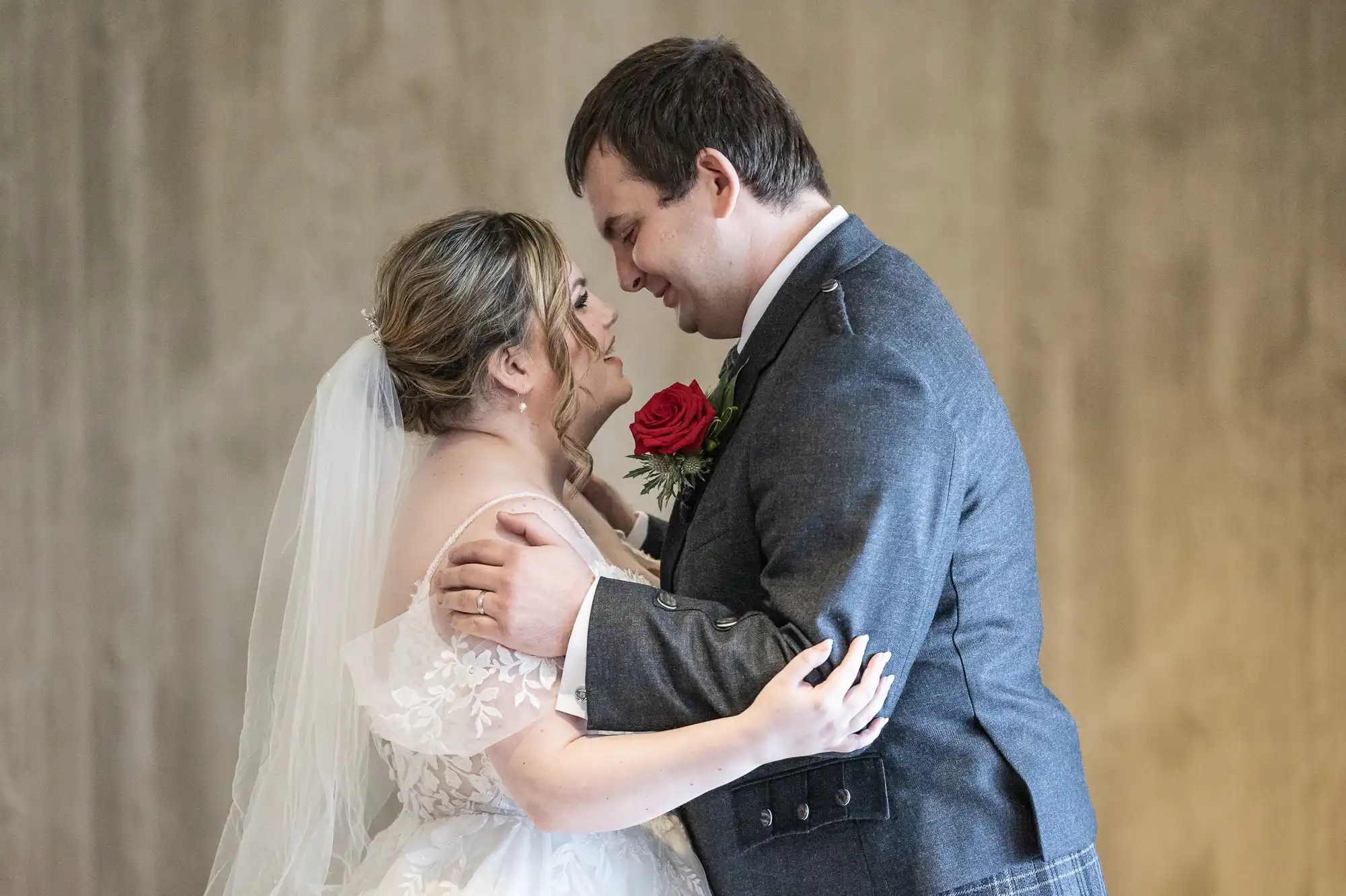 A bride in a white dress and veil smiles as she embraces a groom in a grey suit with a red rose buttonhole. They stand close, touching foreheads and gazing into each other’s eyes against a neutral, blurred background.