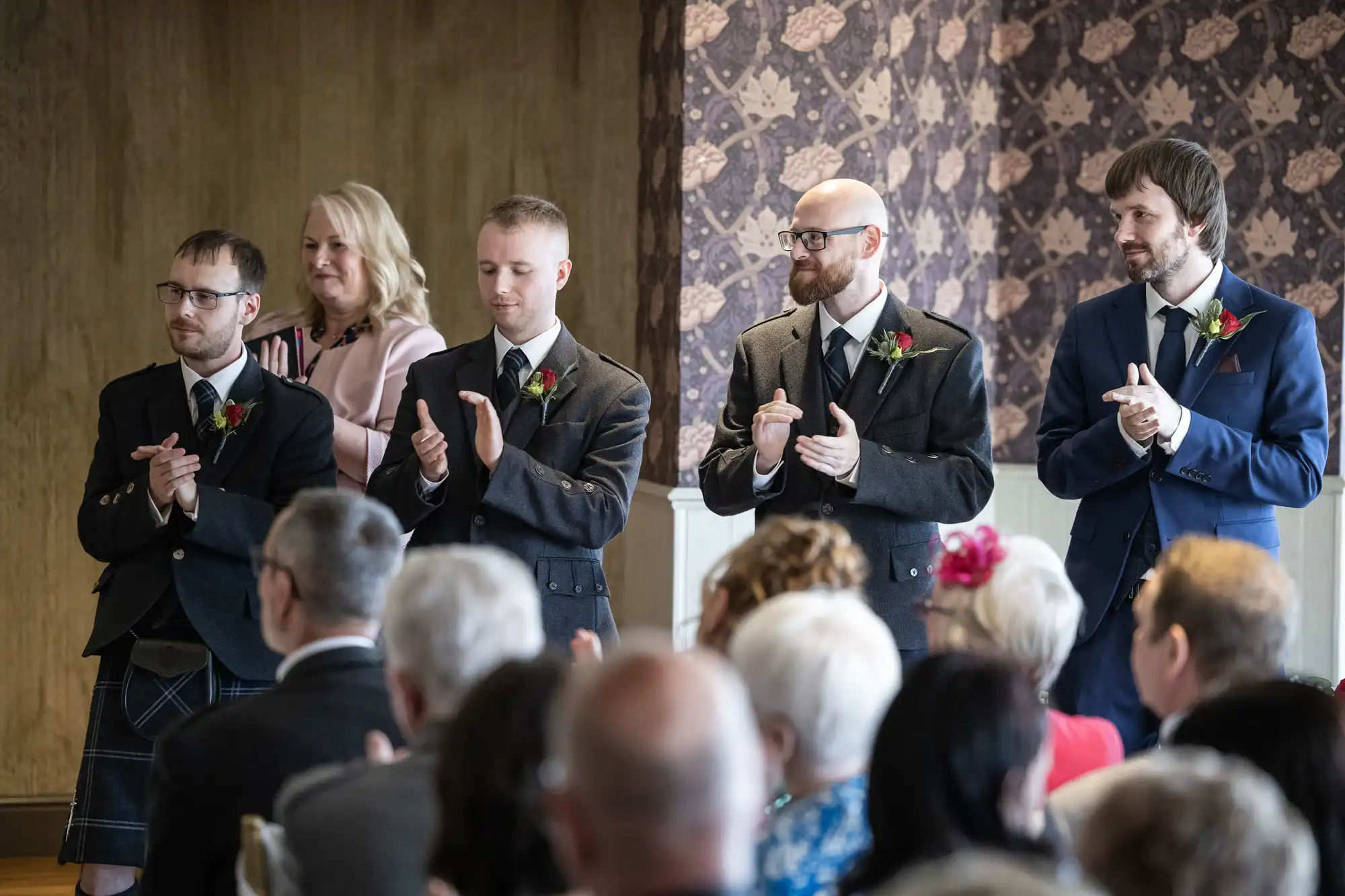 Four men in formal attire and a woman in a pink jacket stand side by side, applauding at an indoor event. The background features patterned wallpaper; several seated guests with their backs to the camera fill the foreground.