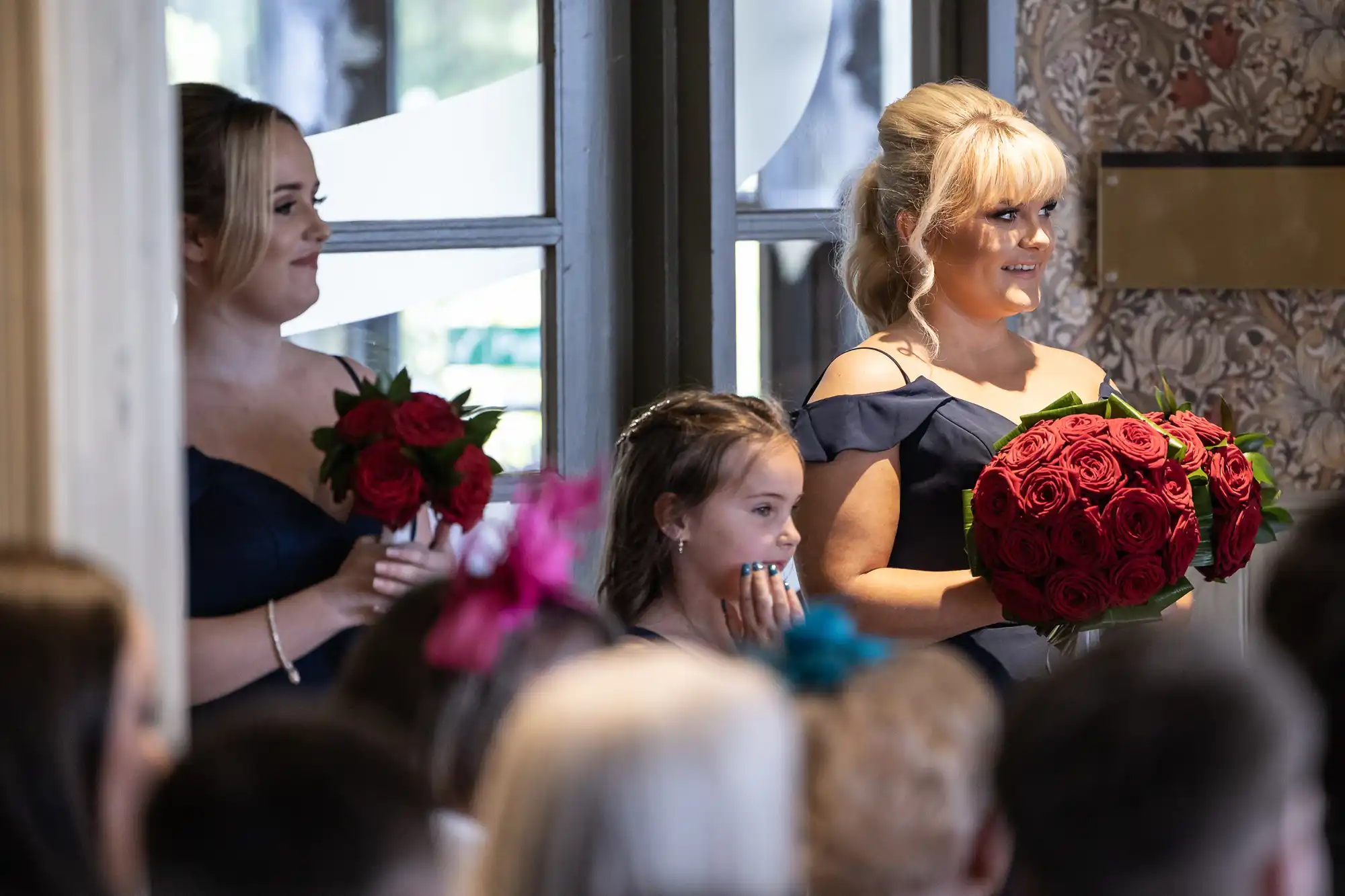 Two women in navy dresses hold red rose bouquets and smile, standing indoors at a formal event. A young girl between them clasps her hands to her face, appearing excited. People with colourful hats and hair are seated in the foreground, backs to the camera.
