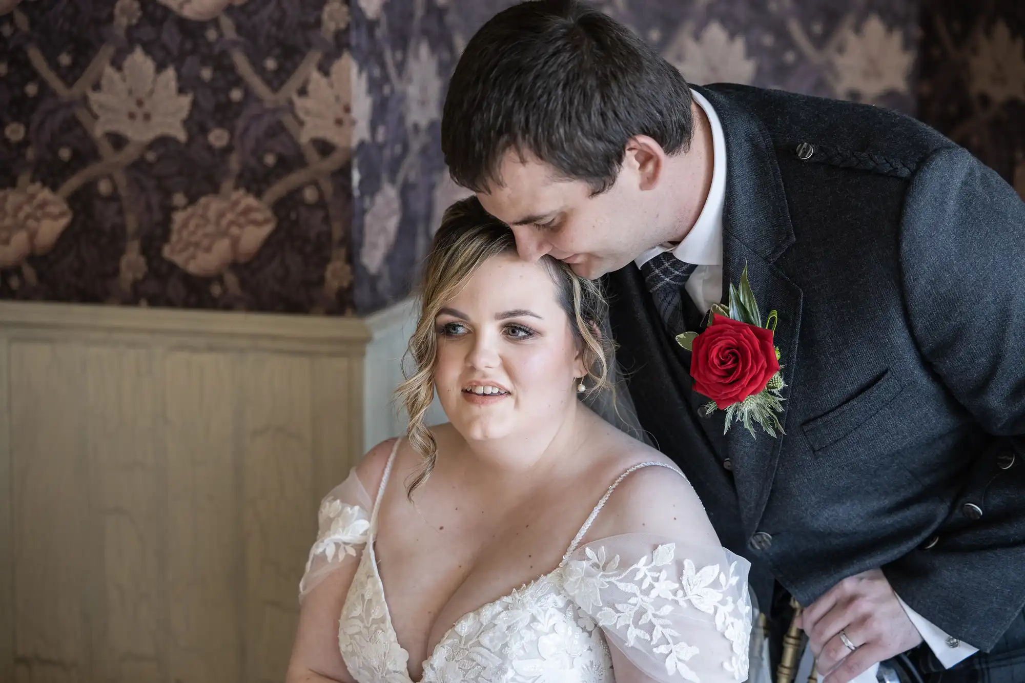 A bride in a white, off-shoulder lace wedding dress sits, smiling softly. A groom in a dark suit with a red rose buttonhole gently leans down to kiss her head. The background features floral-patterned wallpaper and wooden panelling.