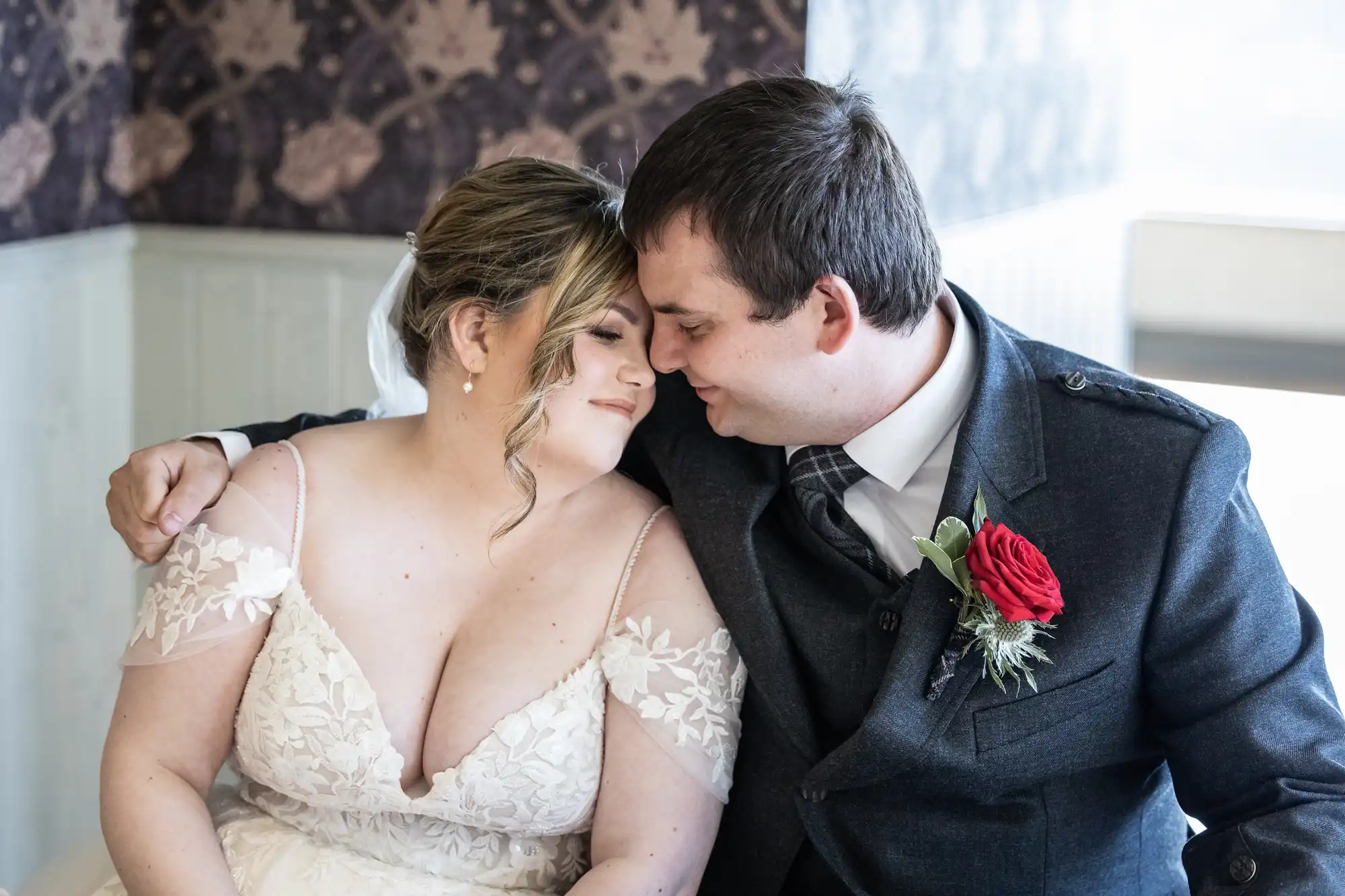 A bride and groom sit close together, touching foreheads and smiling gently. The bride wears an off-the-shoulder white lace gown. The groom wears a dark suit with a red rose buttonhole. They appear happy and content in a softly lit room.