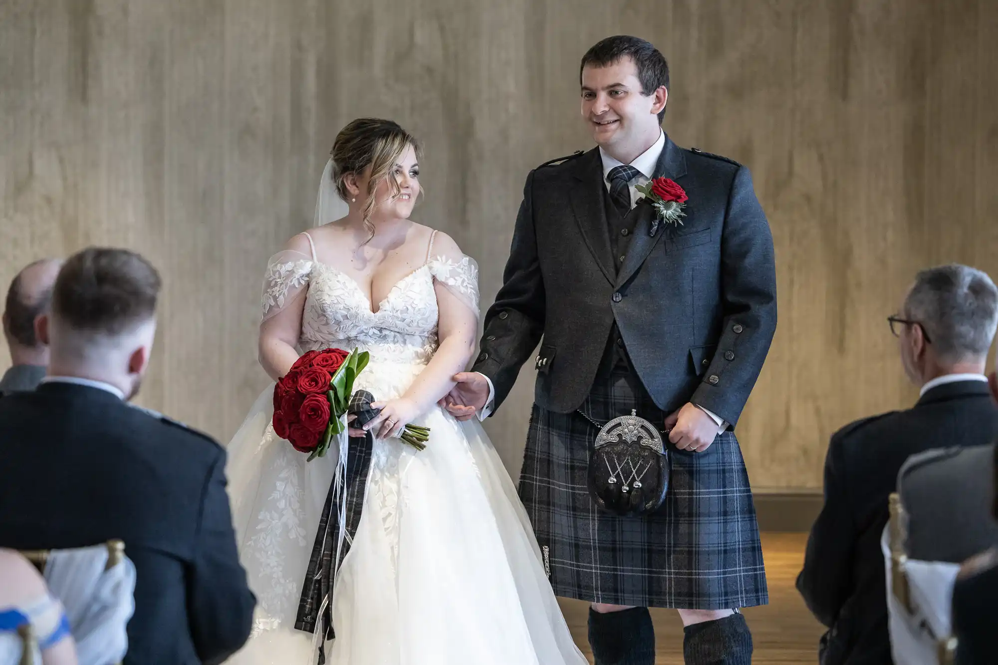 A bride in a white dress holding red roses stands next to a groom in a dark jacket and tartan kilt. They smile at each other during a wedding ceremony, with guests seated facing them. The background is a plain, textured wall.