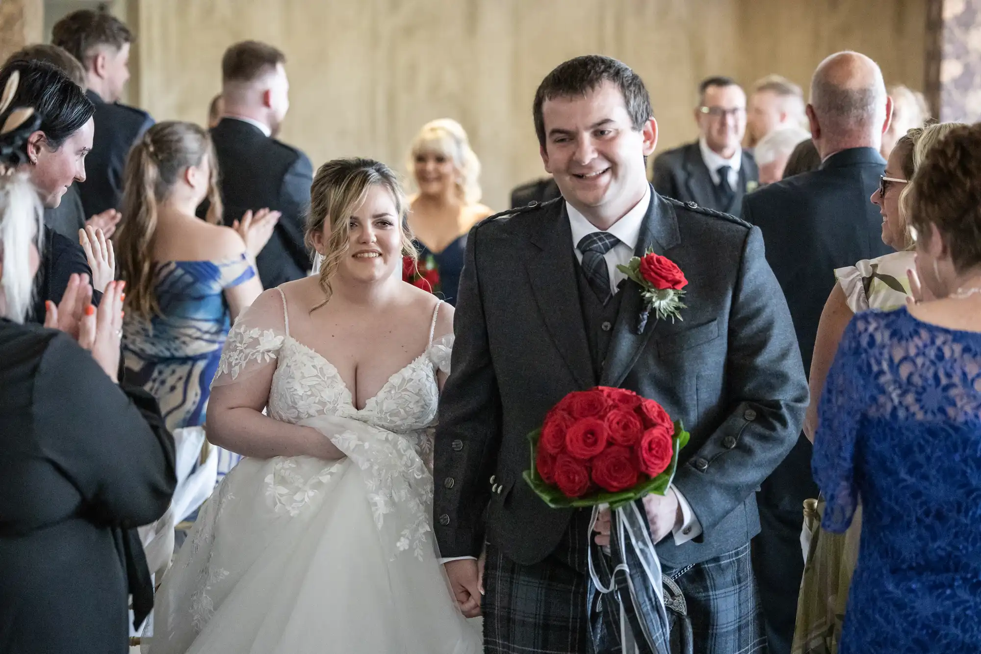 A bride in a white dress and a groom in a dark suit with a kilt smile as they walk down the aisle, holding hands. The groom carries a bunch of red roses. Guests line both sides, clapping and smiling, with blurred background details.