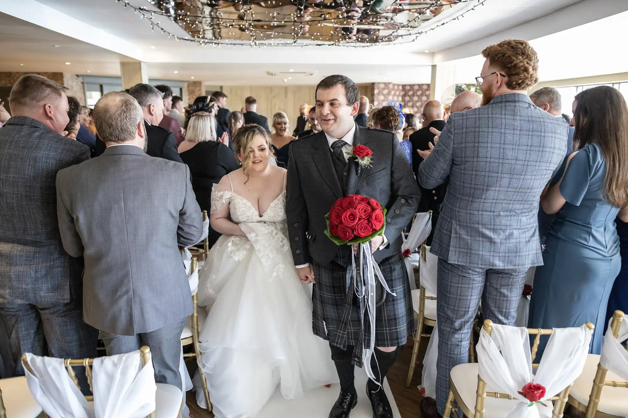 A bride in a white gown and a groom in a kilt holding red roses walk hand-in-hand down the aisle, smiling, whilst wedding guests stand and face them in celebration, filling a bright indoor venue with gold chairs and festive lights overhead.