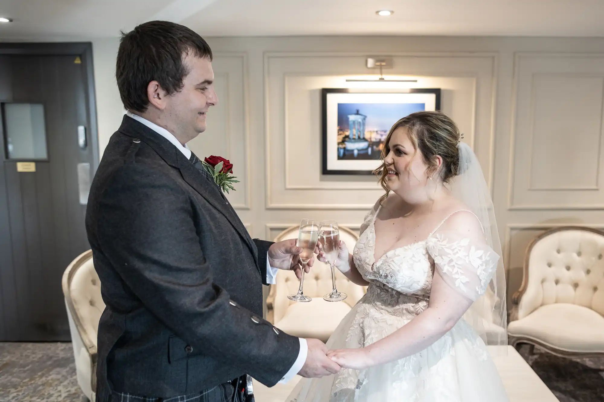 A bride in a white, off-the-shoulder wedding gown and a groom in a dark suit with a red rose buttonhole smile and hold hands while toasting with champagne glasses in a bright, elegant room with cream chairs and framed artwork on the wall behind them.
