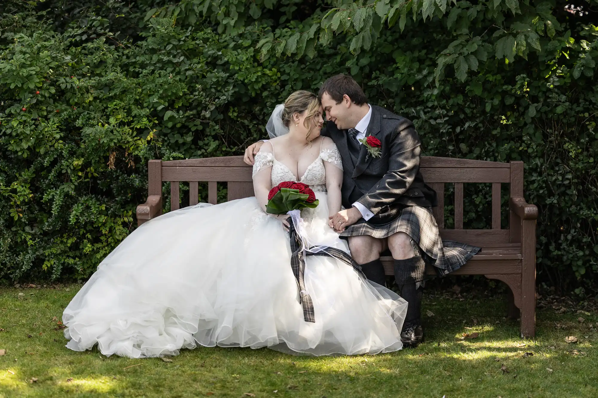 A bride in a white wedding dress and a groom in a Scottish kilt sit closely on a wooden bench, touching foreheads and smiling. The bride holds a bouquet of red roses. Lush green foliage fills the background.