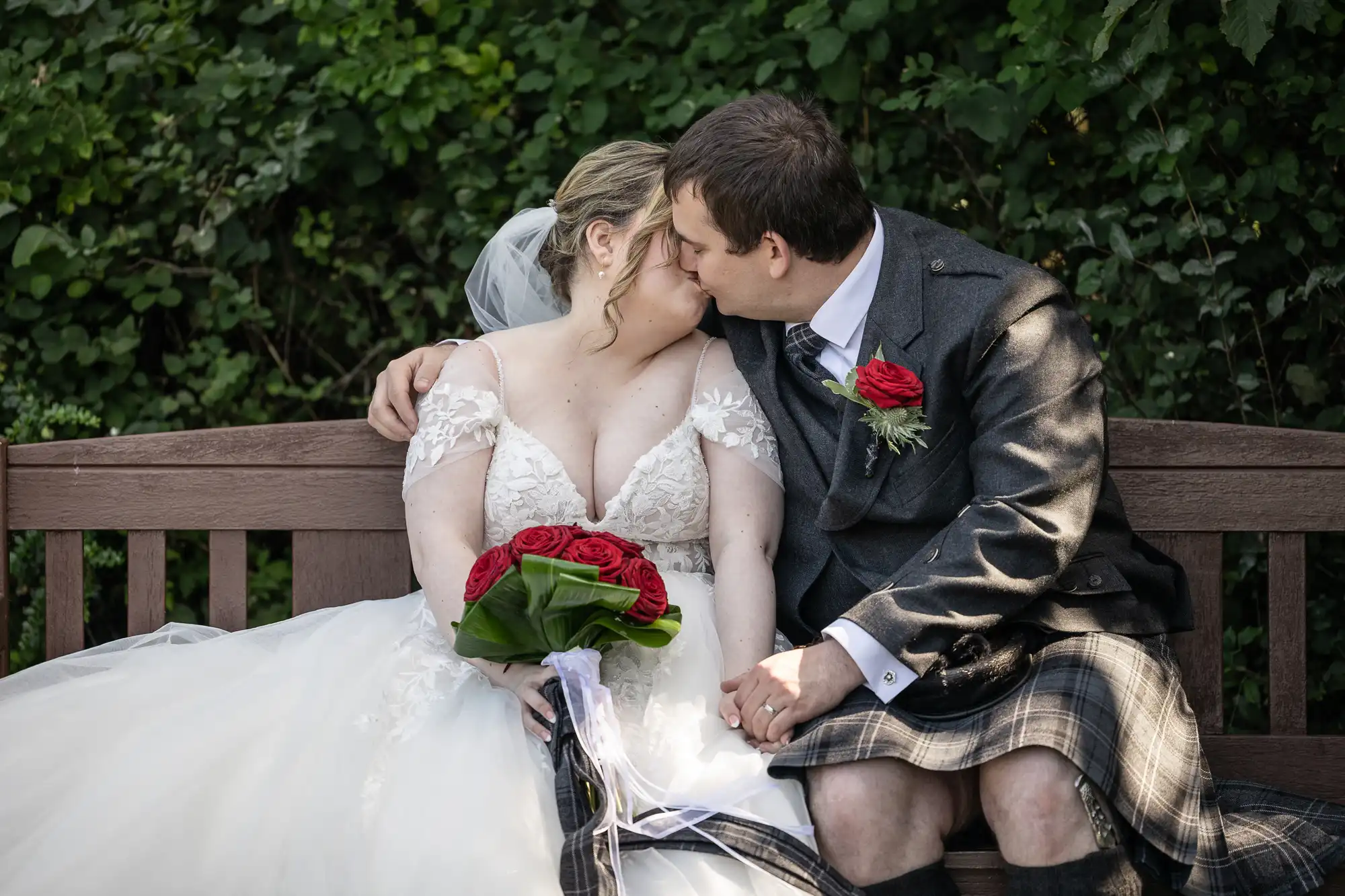 A bride in a white gown and a groom in a grey jacket and plaid kilt sit on a wooden bench, holding hands and kissing. The bride holds a bouquet of red roses, and both wear red rose buttonholes. Lush green foliage forms the background.