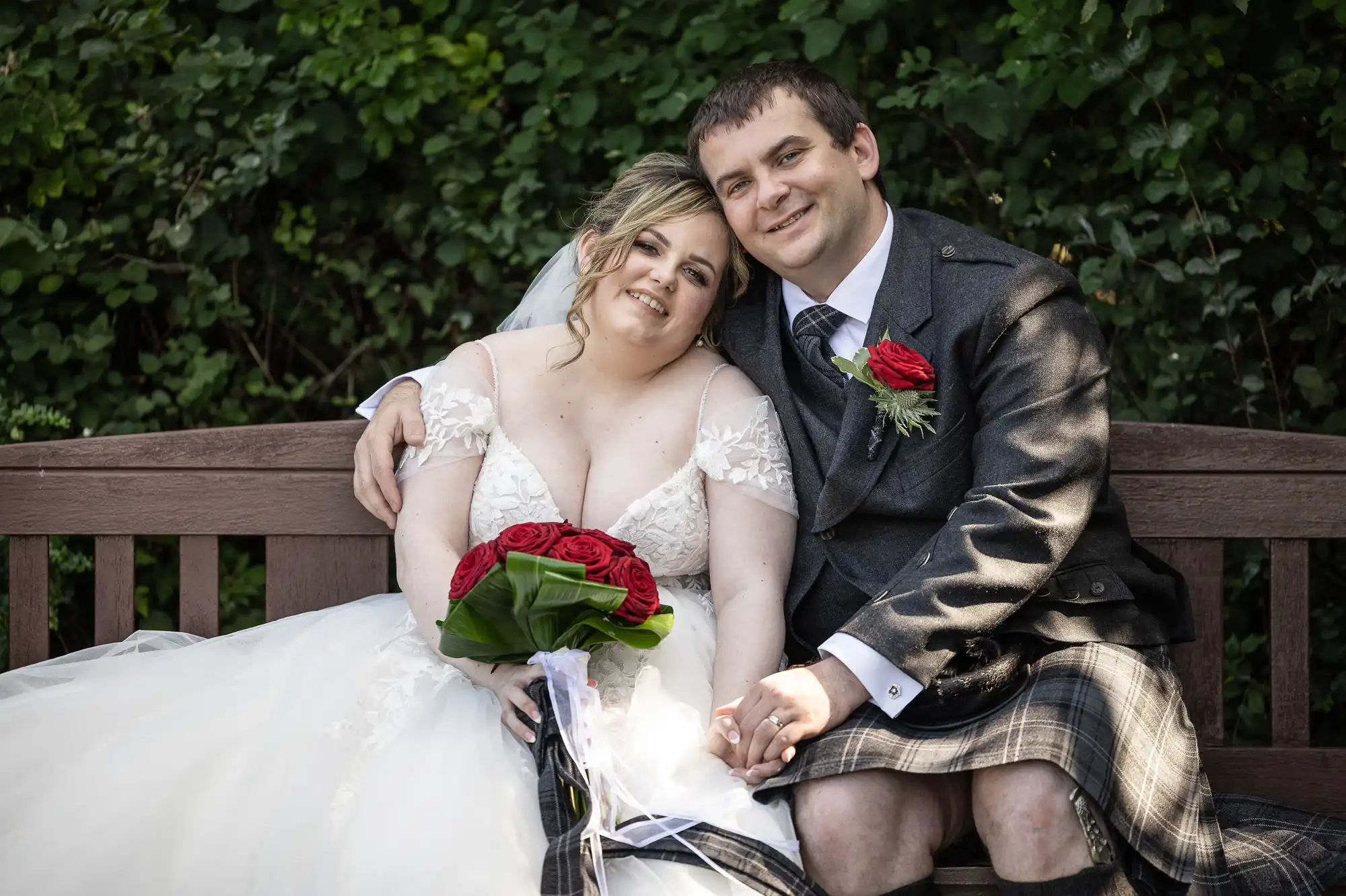 A smiling bride in a white dress holds red roses and leans on a groom in a kilt and jacket. They sit close together on a wooden bench, holding hands, with green foliage in the background. Both look happy and relaxed.