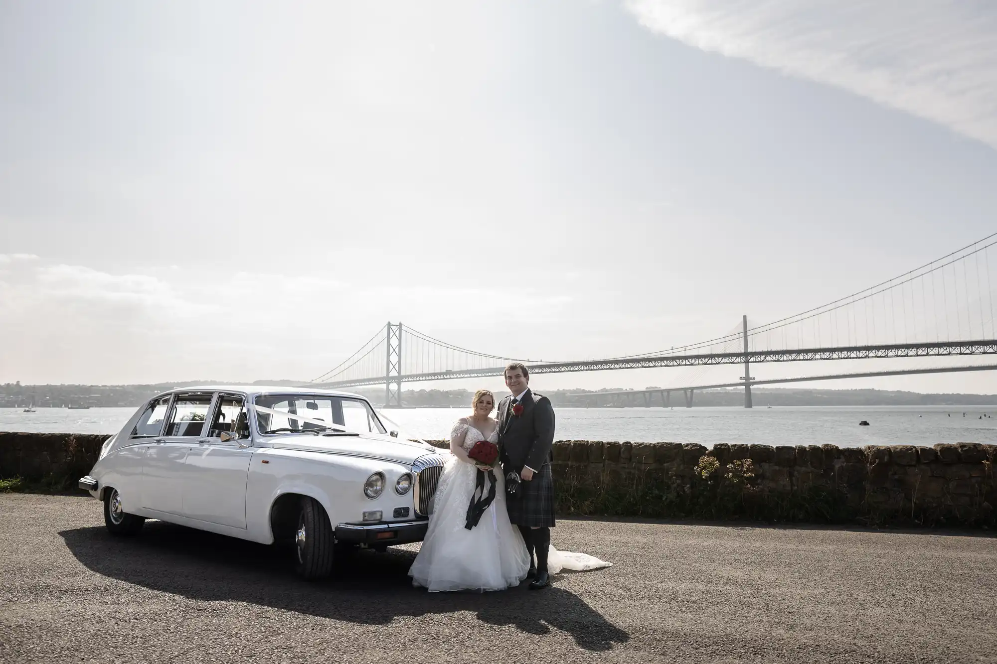 A bride and groom stand together beside a vintage white car near a stone wall, with a large suspension bridge and water in the background under a bright, partly cloudy sky. The bride wears a white dress and holds a bouquet; the groom wears a grey suit with a kilt.