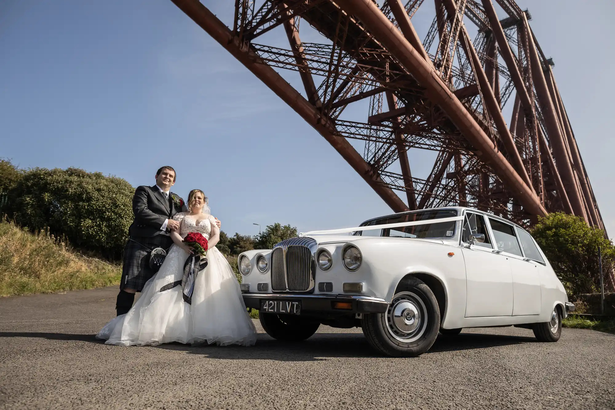 A bride in a white gown and groom in a kilt stand smiling beside a classic white car under a large red steel bridge on a sunny day, with greenery in the background. The bride holds a bouquet of red and white flowers.
