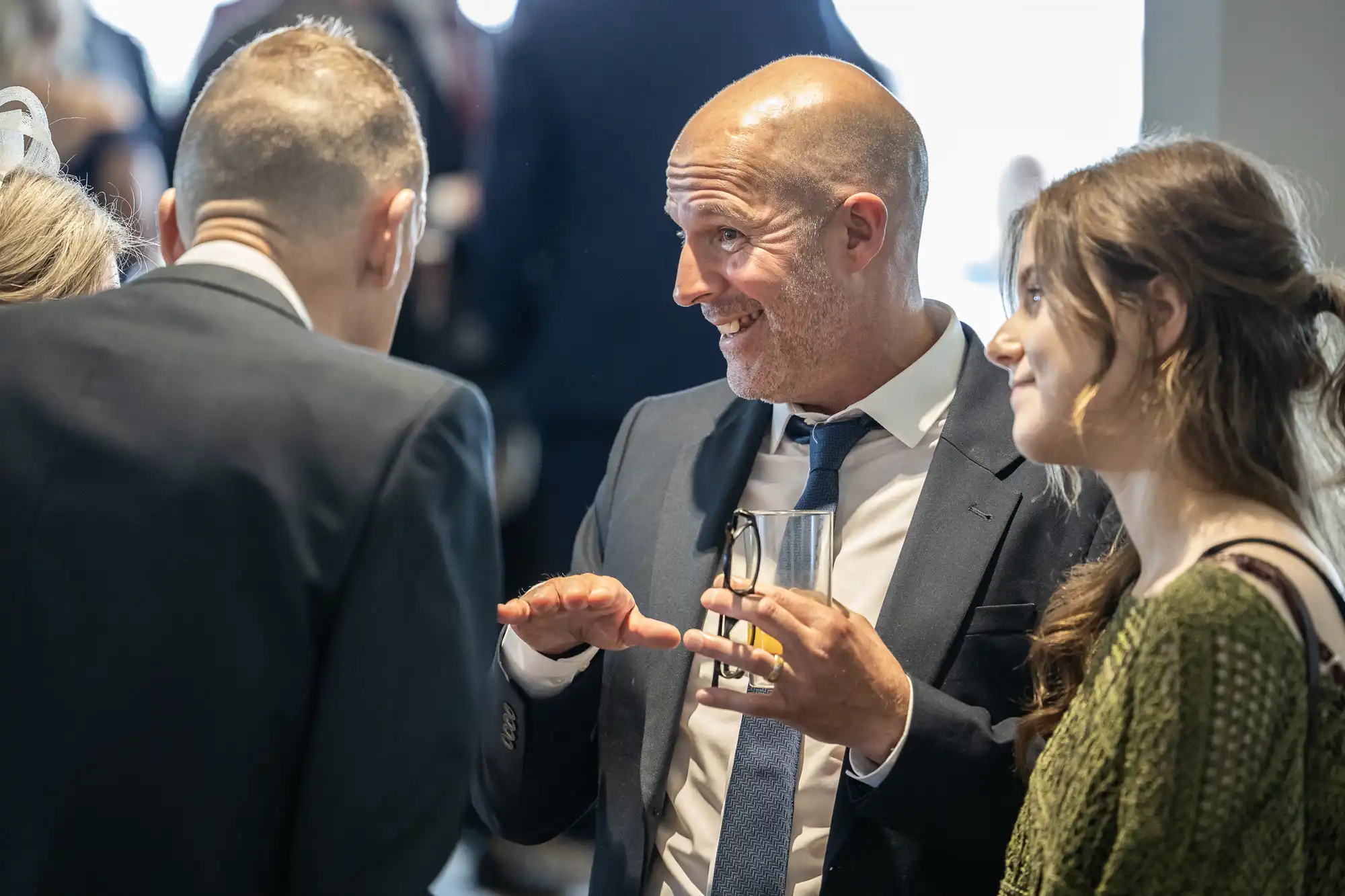 A bald man in a suit and tie gestures animatedly while holding a glass, talking to a man in a suit with his back to the camera and a smiling woman with long brown hair in a green jumper. They are indoors, possibly at a social or professional event.