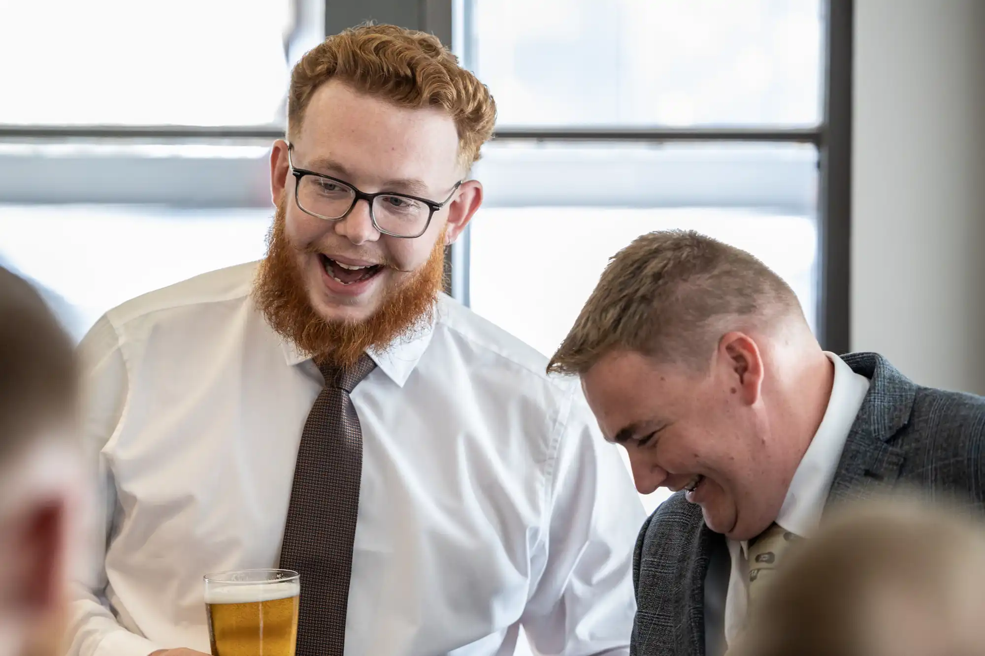 Two men dressed in business attire laugh together indoors. The man on the left, with red hair, glasses, and a beard, holds a pint of beer and smiles widely. The man on the right, with short hair, looks down whilst laughing. Both appear to be enjoying a light-hearted moment.