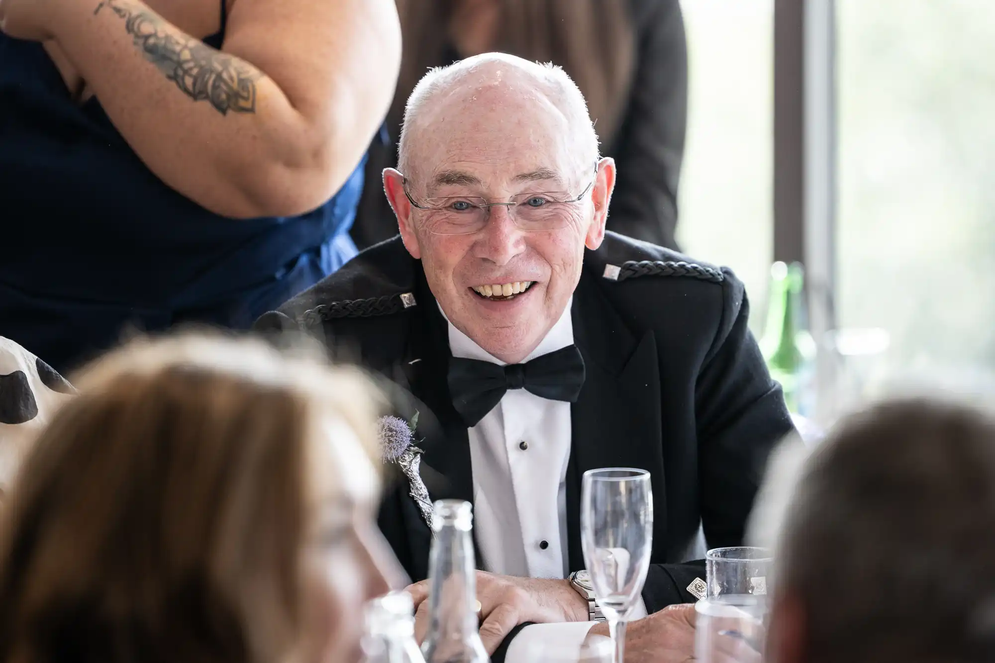 An older man with glasses, wearing a black dinner jacket and bow tie, smiles while seated at a table set with empty champagne glasses and bottles. Other people are blurred in the foreground, and a tattooed arm is visible in the background.