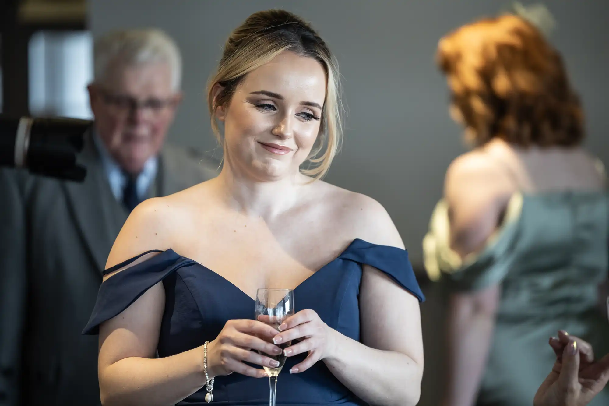 A woman in an off-the-shoulder navy dress holds a champagne flute and smiles softly. Behind her, an older man in a suit and tie stands blurred, whilst another woman in a green dress is turned away. The setting appears to be a formal event or celebration.