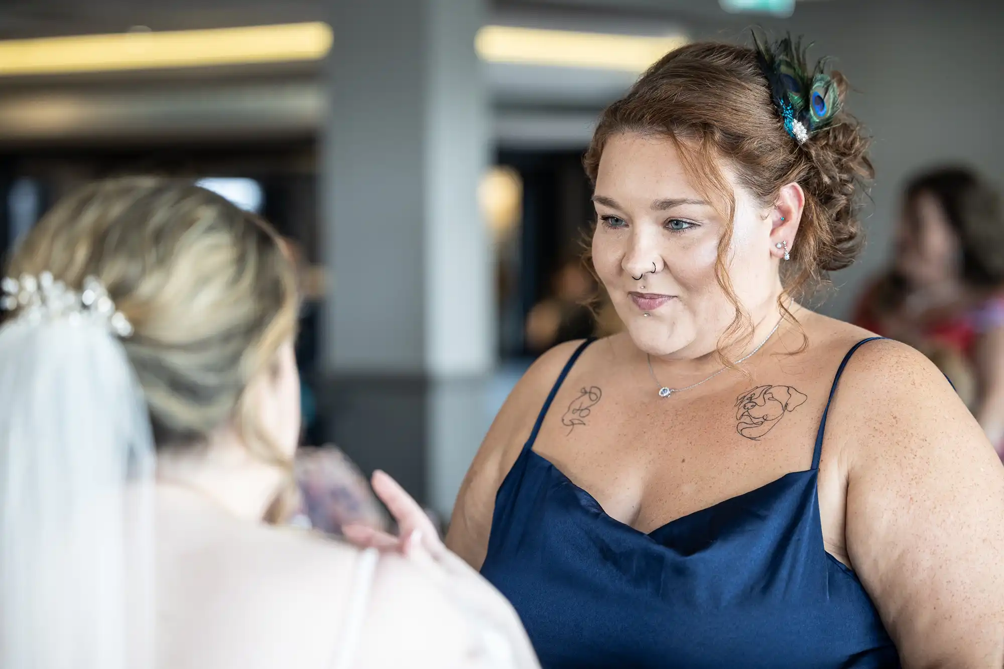 A woman in a navy blue dress with shoulder tattoos and a peacock feather in her hair smiles at another woman in a wedding dress and veil. The background is softly blurred, suggesting an indoor event or celebration.