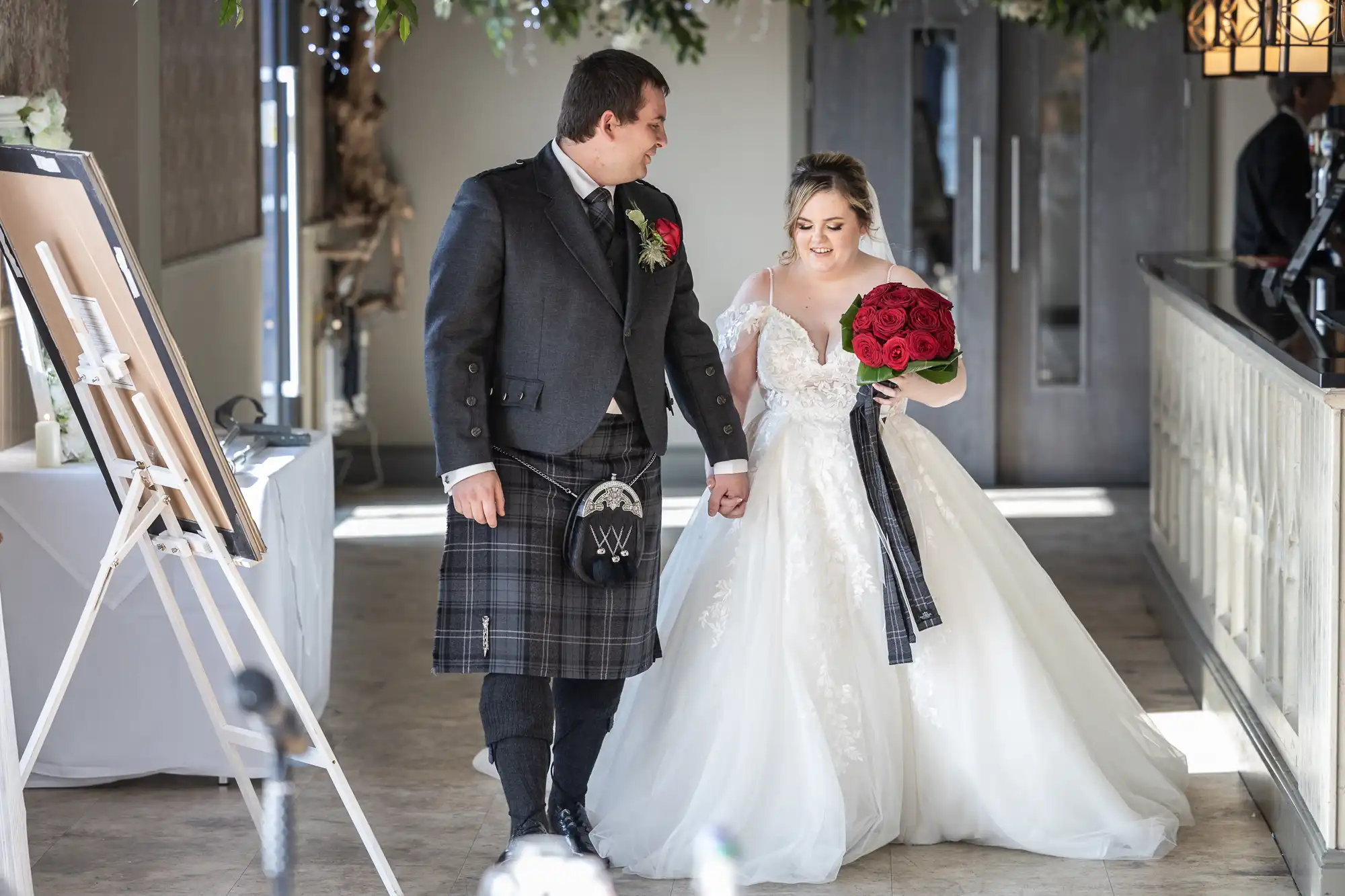 A groom in a grey jacket and tartan kilt holds hands with a bride in a white lace gown and veil. The bride carries a bouquet of red roses with a tartan ribbon. They smile at each other while walking indoors, under hanging greenery and soft lighting.