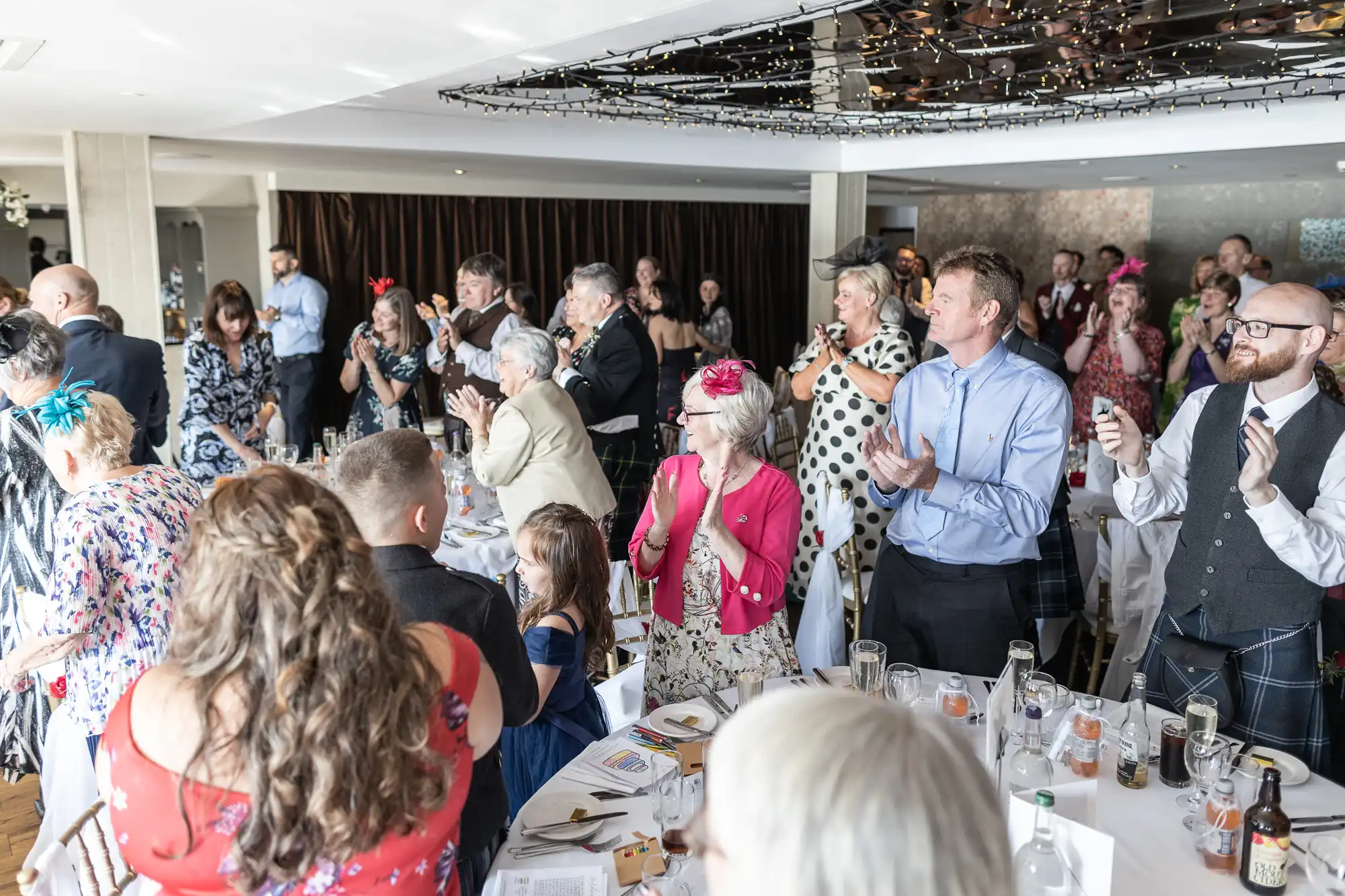 A large group of people dressed in formal attire stand and applaud at decorated round tables in a bright, modern event space. The atmosphere is celebratory, with drinks, floral hats, and table settings visible, suggesting a wedding or formal gathering.