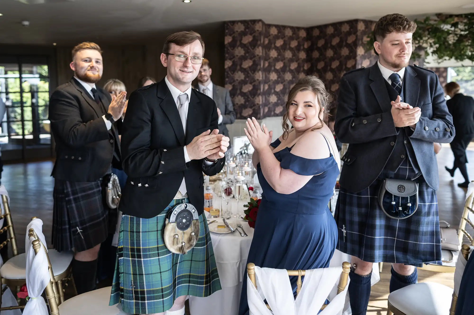 A group of five people in formal attire, including kilts and jackets, stand and clap around a decorated table at an indoor event. One woman in a navy dress smiles at the camera; the men wear kilts in various tartans. The background shows windows and patterned walls.