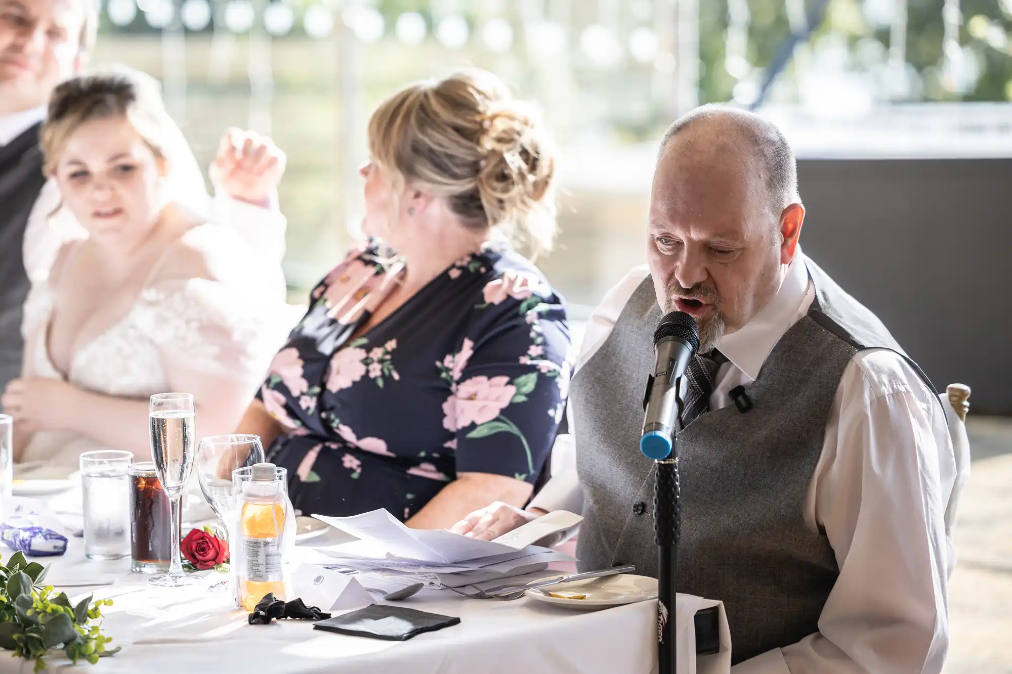 A man in a grey waistcoat speaks into a microphone whilst reading papers at a wedding reception table. A woman in a floral dress and another woman in a white dress sit beside him, listening. Sunlight streams in from large windows behind them.