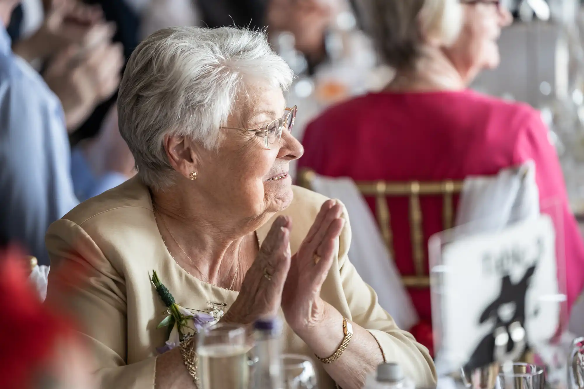 An elderly woman with short grey hair and glasses smiles and claps whilst seated at a table during a formal event. She wears a beige jacket with a corsage. Blurred guests in colourful attire and glassware fill the bright, festive background.