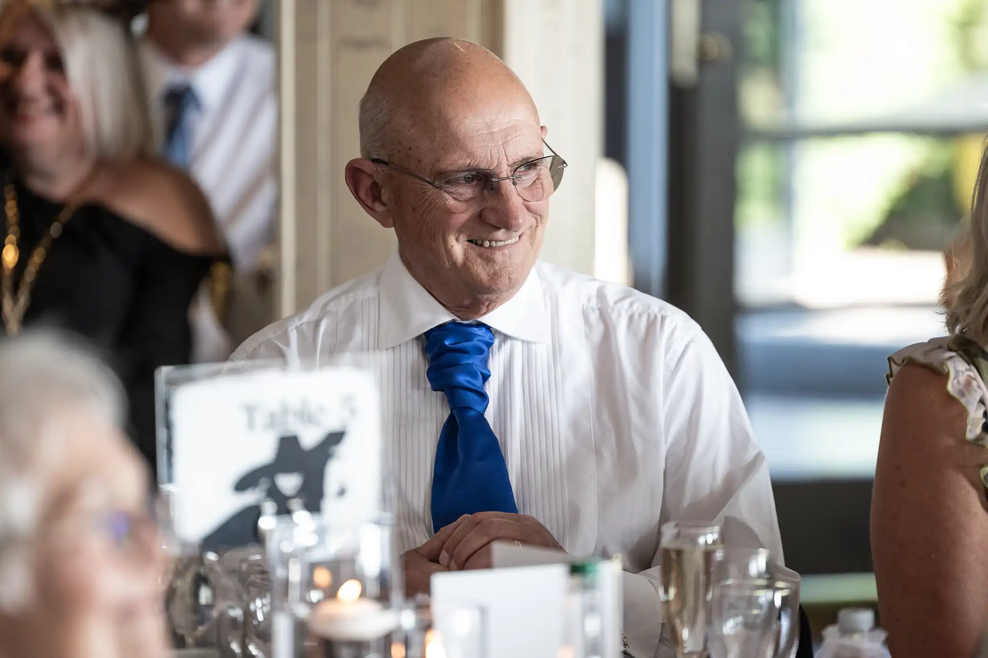 An older man with glasses and a bright blue tie sits at a table, smiling. He wears a white shirt and is surrounded by other people. The table has glasses, candles, and a sign reading "Table 5." The background is softly blurred.