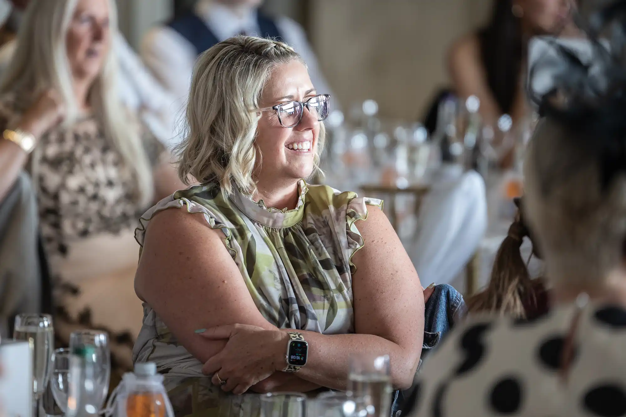 A woman with blonde, wavy hair and glasses smiles whilst seated at a table during an event. She wears a patterned sleeveless top and an Apple Watch. Blurred people and drinking glasses are visible in the background.
