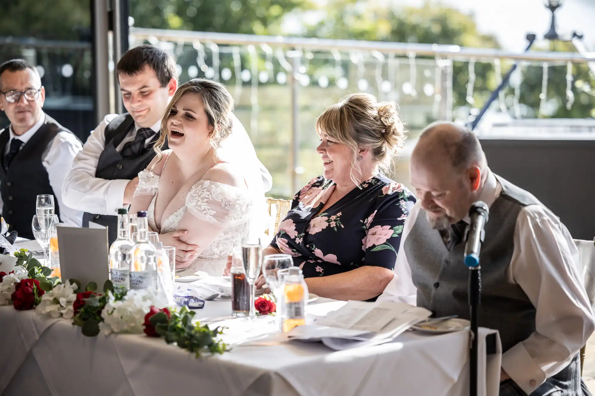 A bride laughs beside the groom and a woman in a floral dress at a decorated wedding reception table. A man at the microphone reads from papers, whilst another man sits at the end. Sunlight streams in from large windows behind them.