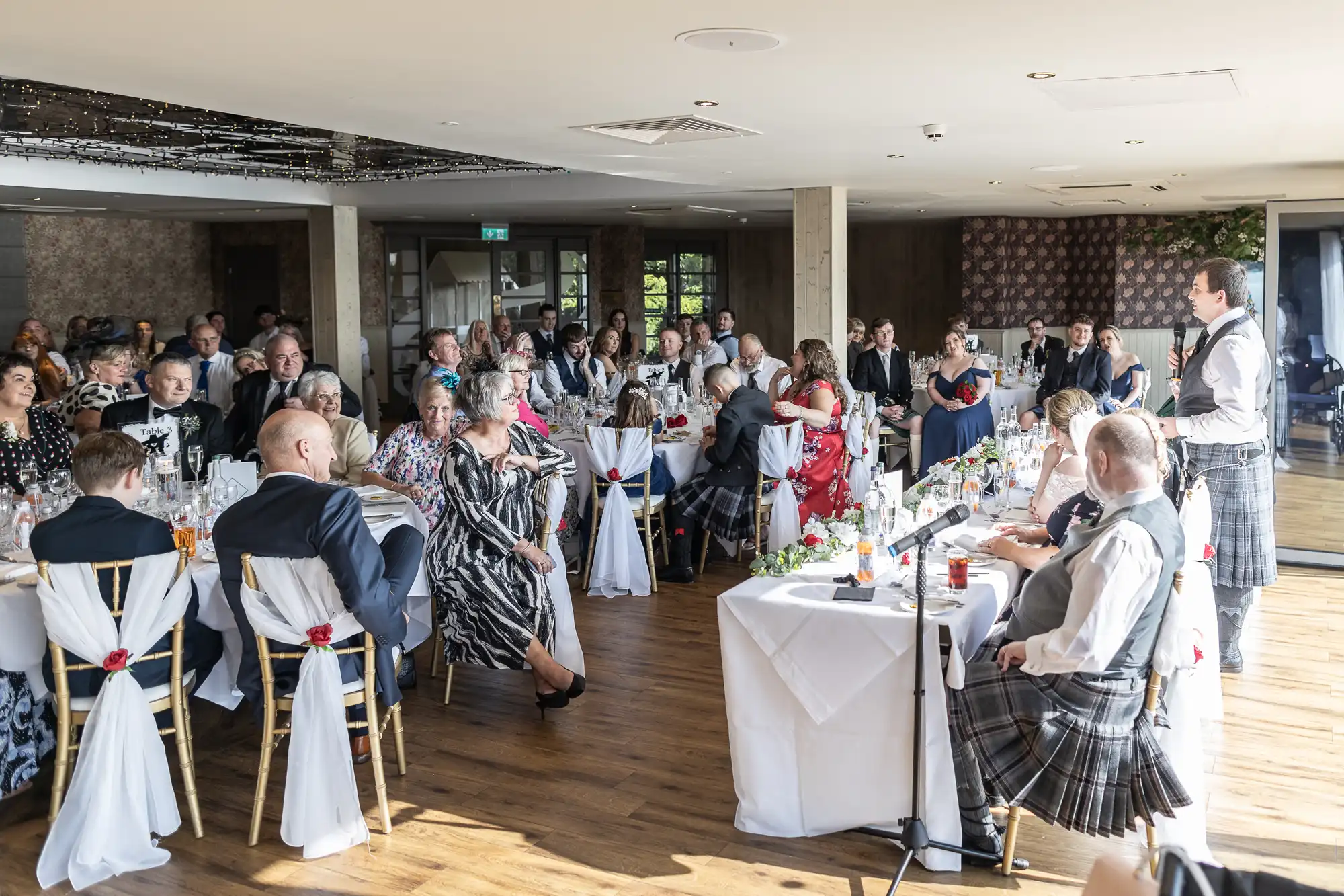 A wedding reception in a bright room with wooden floors. Guests in formal attire sit at decorated tables, listening to a man in a kilt giving a speech at the top table. White tablecloths, floral centrepieces, and sunlight fill the lively scene.